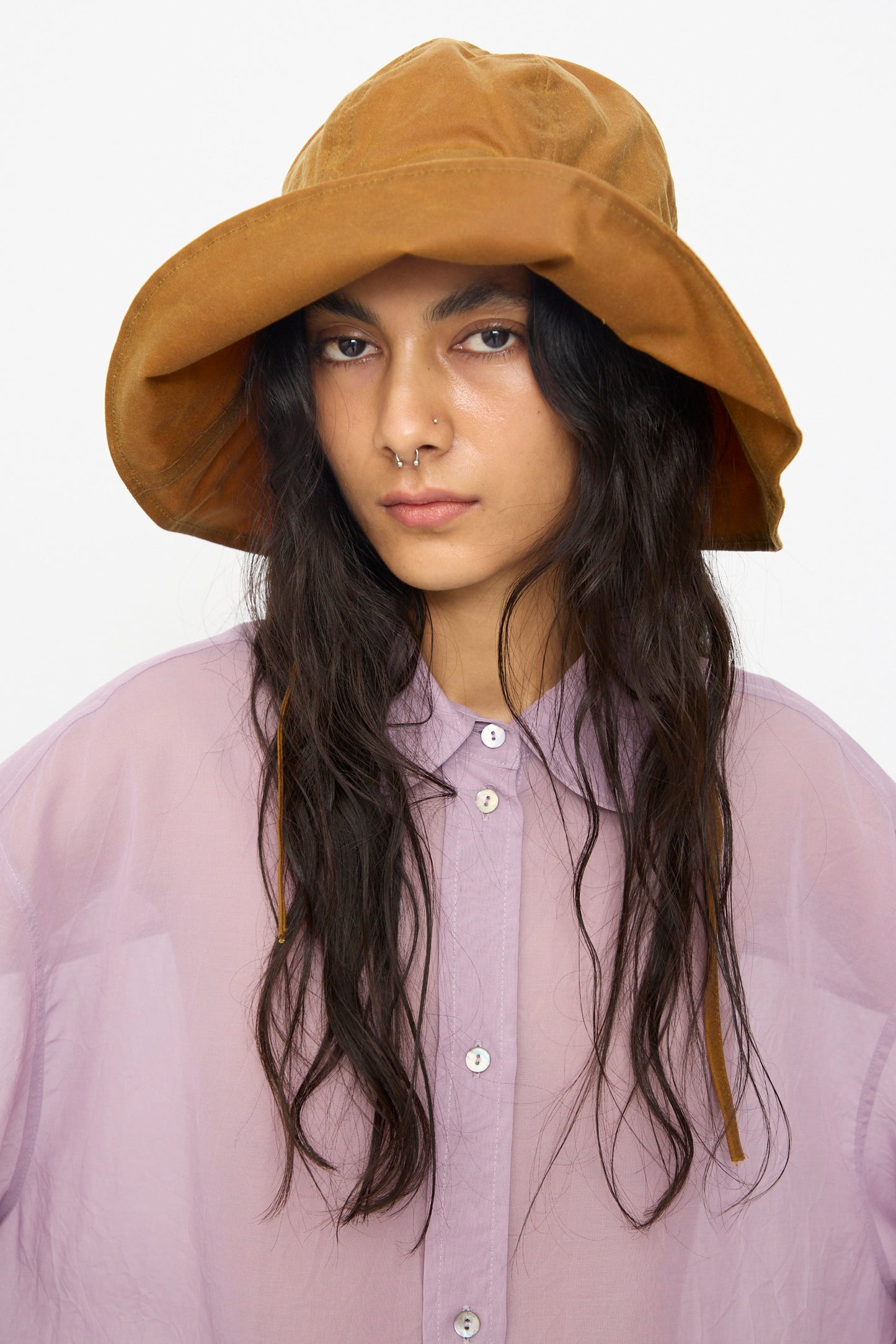 A person wears the Cawley British Waxed Oilskin Sunhat in Gold and a sheer lavender button-up shirt, looking at the camera with a neutral expression.