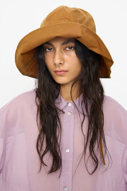 A person wears the Cawley British Waxed Oilskin Sunhat in Gold and a sheer lavender button-up shirt, looking at the camera with a neutral expression.