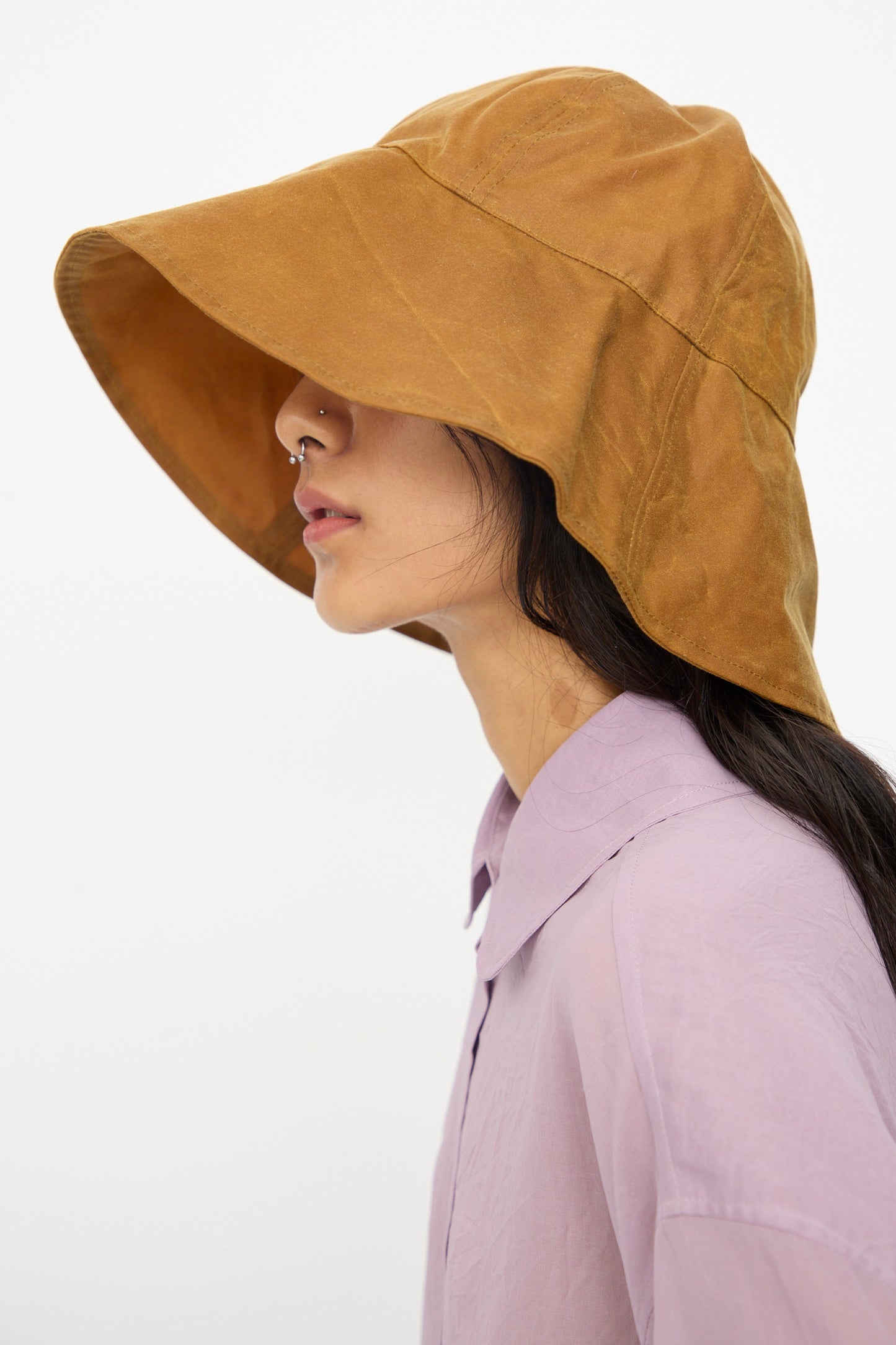 A person in a light purple collared shirt and nose ring is seen in profile wearing the Cawley British Waxed Oilskin Sunhat in Gold, set against a plain background.