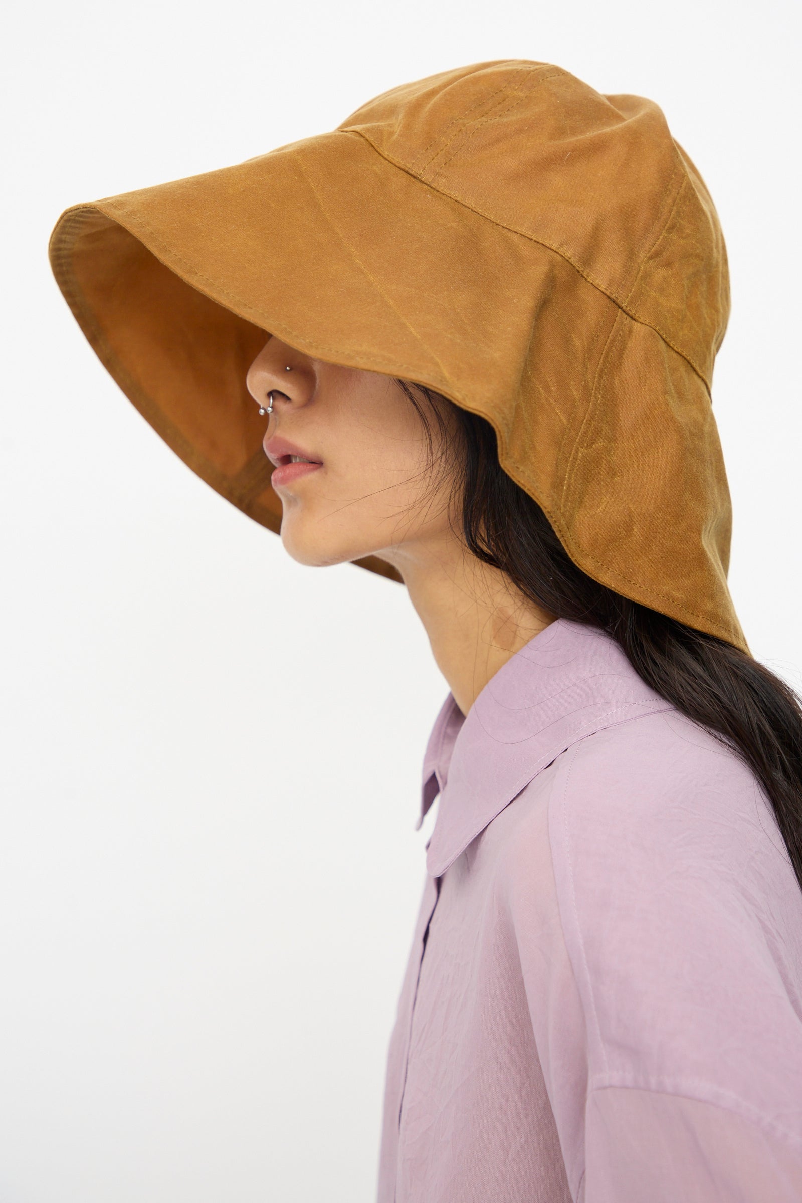 A person in a light purple collared shirt and nose ring is seen in profile wearing the Cawley British Waxed Oilskin Sunhat in Gold, set against a plain background.