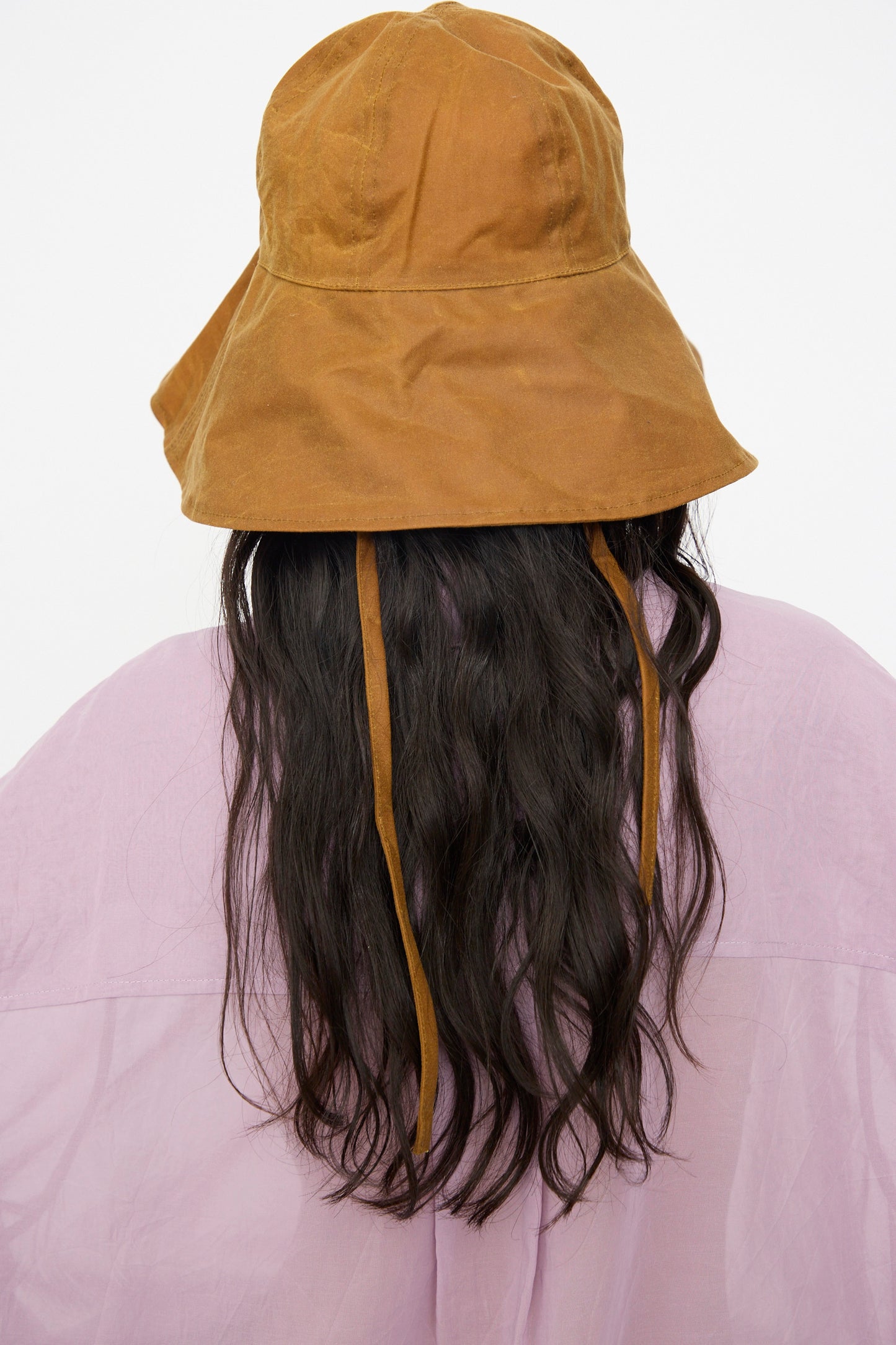 A person, shown from the back with long wavy dark hair, wears a light pink shirt and the Cawley British Waxed Oilskin Sunhat in Gold.