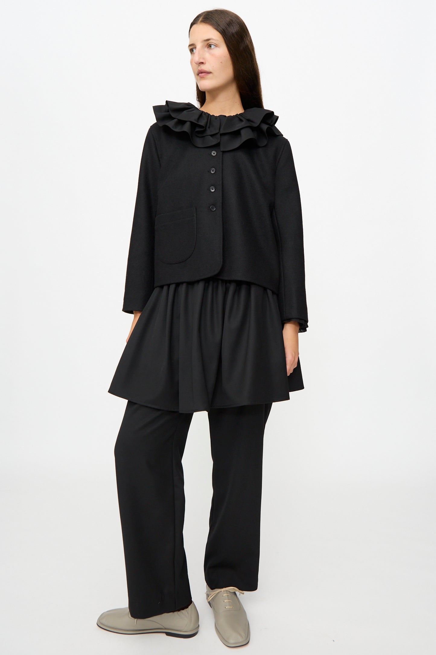 A person stands against a plain white background wearing the Cawley British Wool Rowan Jacket in Black, featuring a ruffled collar and cropped silhouette, styled with a black tunic, loose black pants, and beige lace-up shoes.