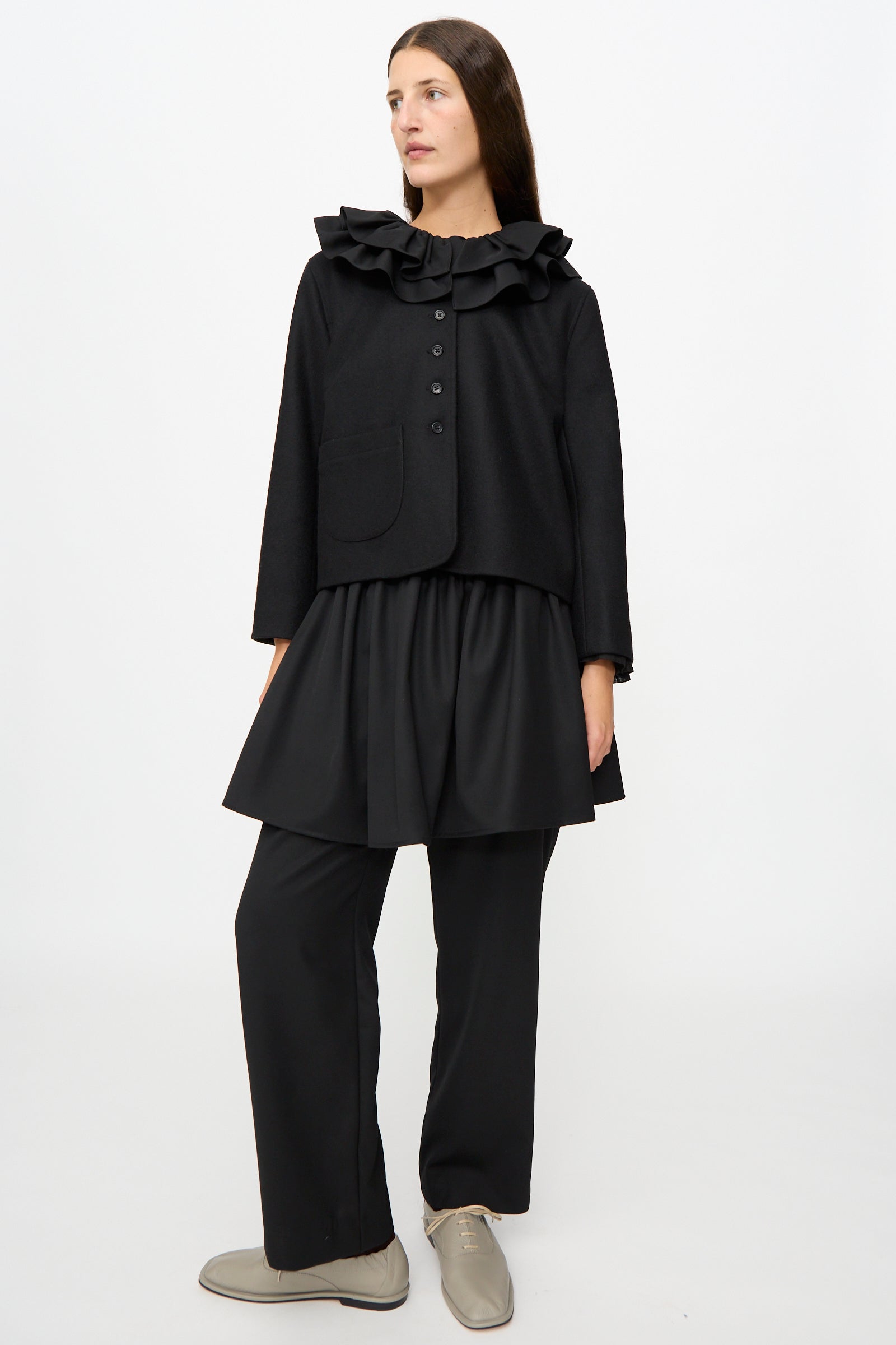 A person stands against a plain white background wearing the Cawley British Wool Rowan Jacket in Black, featuring a ruffled collar and cropped silhouette, styled with a black tunic, loose black pants, and beige lace-up shoes.