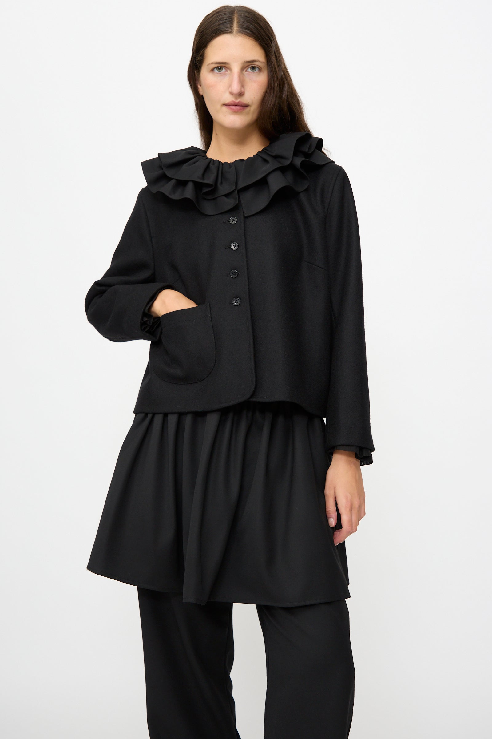 A woman wearing the Cawley British Wool Rowan Jacket in Black, featuring a cropped silhouette and ruffled collar, paired with a black skirt and black pants, stands against a plain white background. This elegant jacket is crafted in the UK from fine British wool.