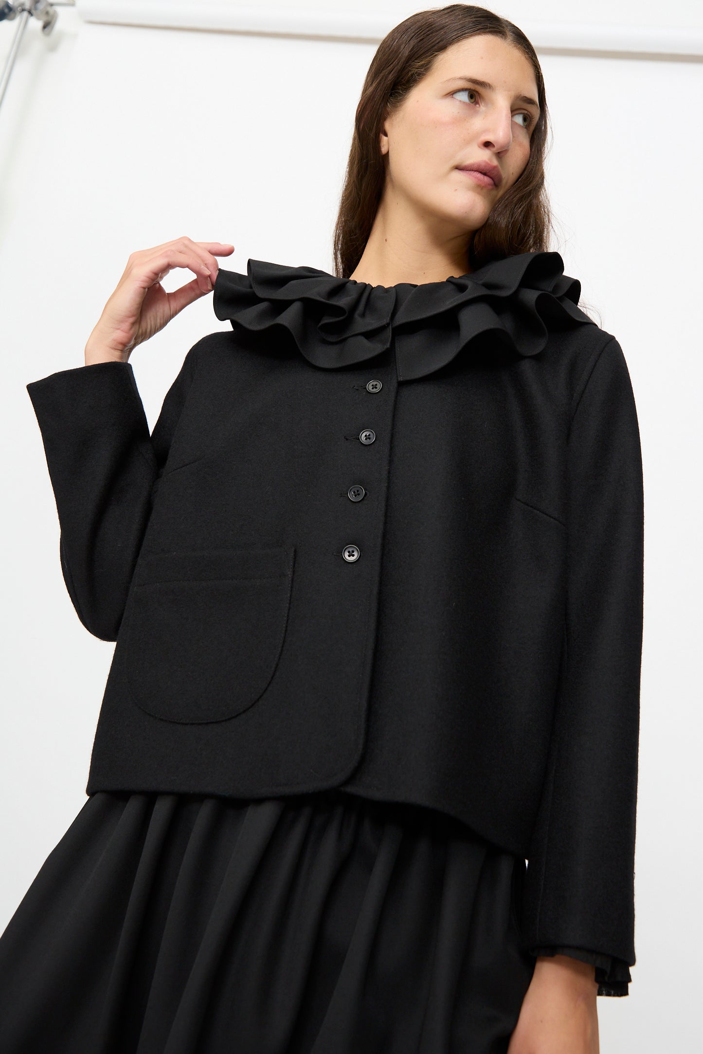 A woman wearing the Cawley British Wool Rowan Jacket in Black—expertly crafted from British wool in the UK with a ruffled collar—and a matching black skirt, stands against a plain white background.