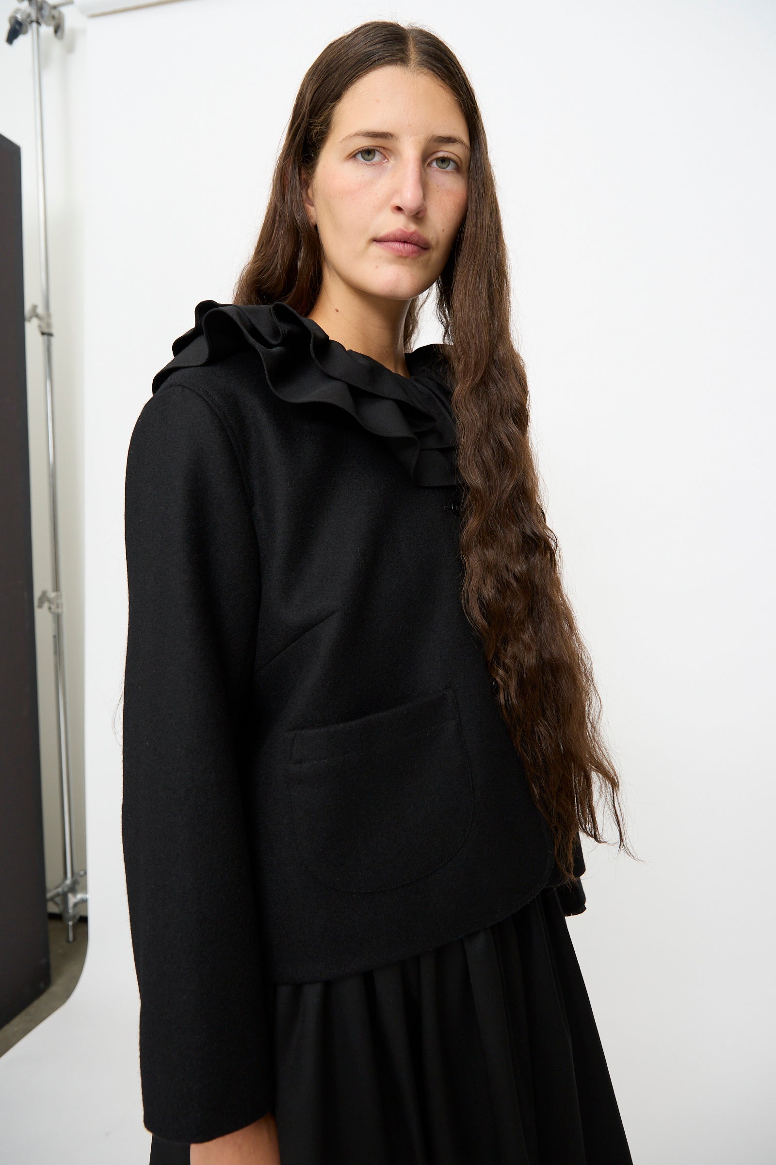 A woman with long brown hair is wearing a black top with a ruffled collar and front pocket, paired with the Cawley British Wool Rowan Jacket in Black, standing against a plain white background.