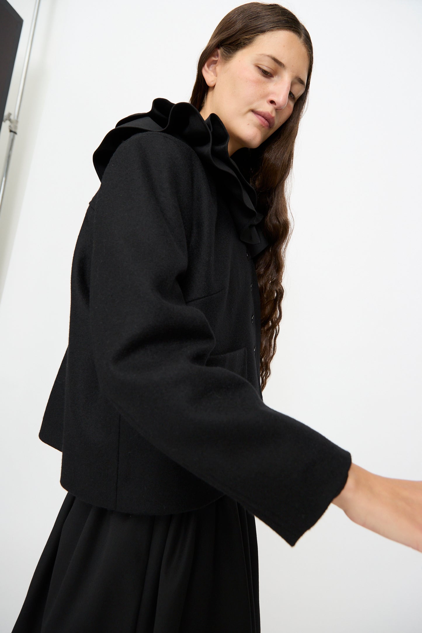 A person with long brown hair wears the Cawley British Wool Rowan Jacket in Black—a cropped style featuring a ruffled collar, expertly crafted from premium British wool and made in the UK—paired elegantly with a matching black skirt, and stands against a plain white background.