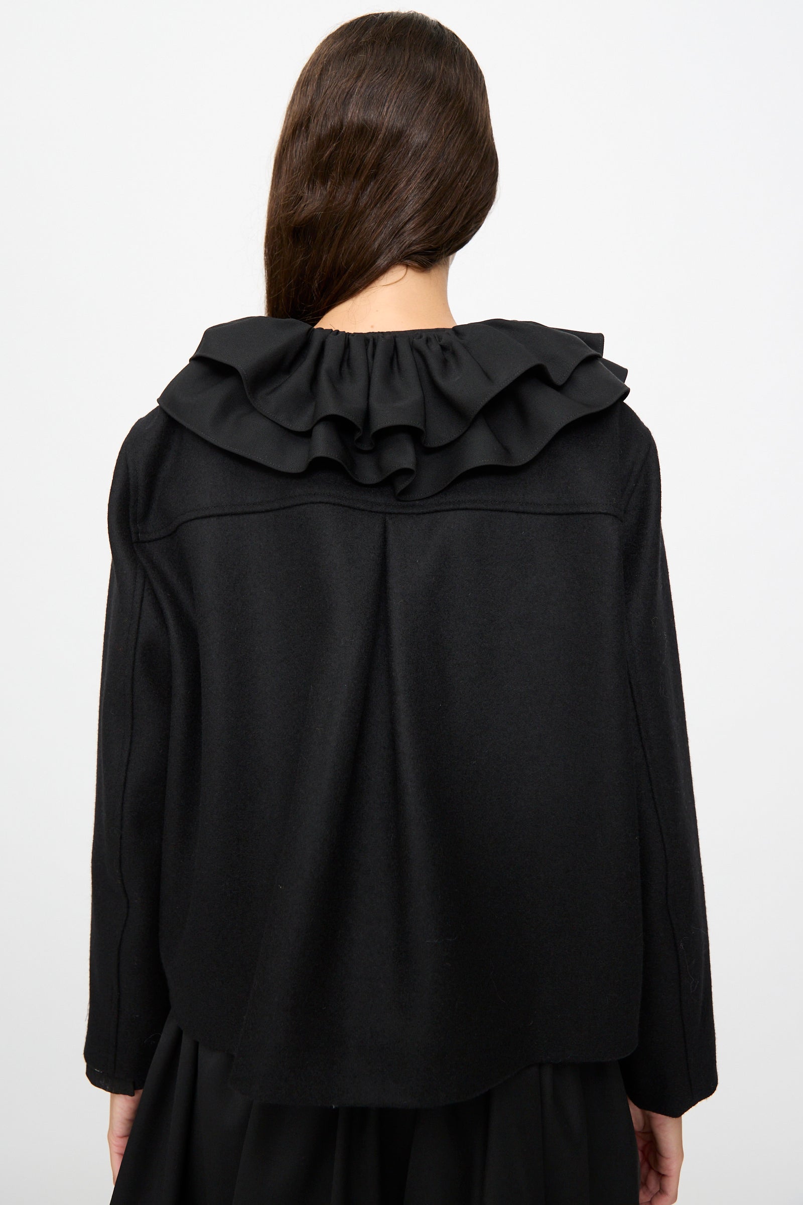 A person with long brown hair, seen from behind, wears the Cawley British Wool Rowan Jacket in Black—a cropped jacket featuring a large ruffled collar and crafted in the UK from fine British wool—set against a plain light background.