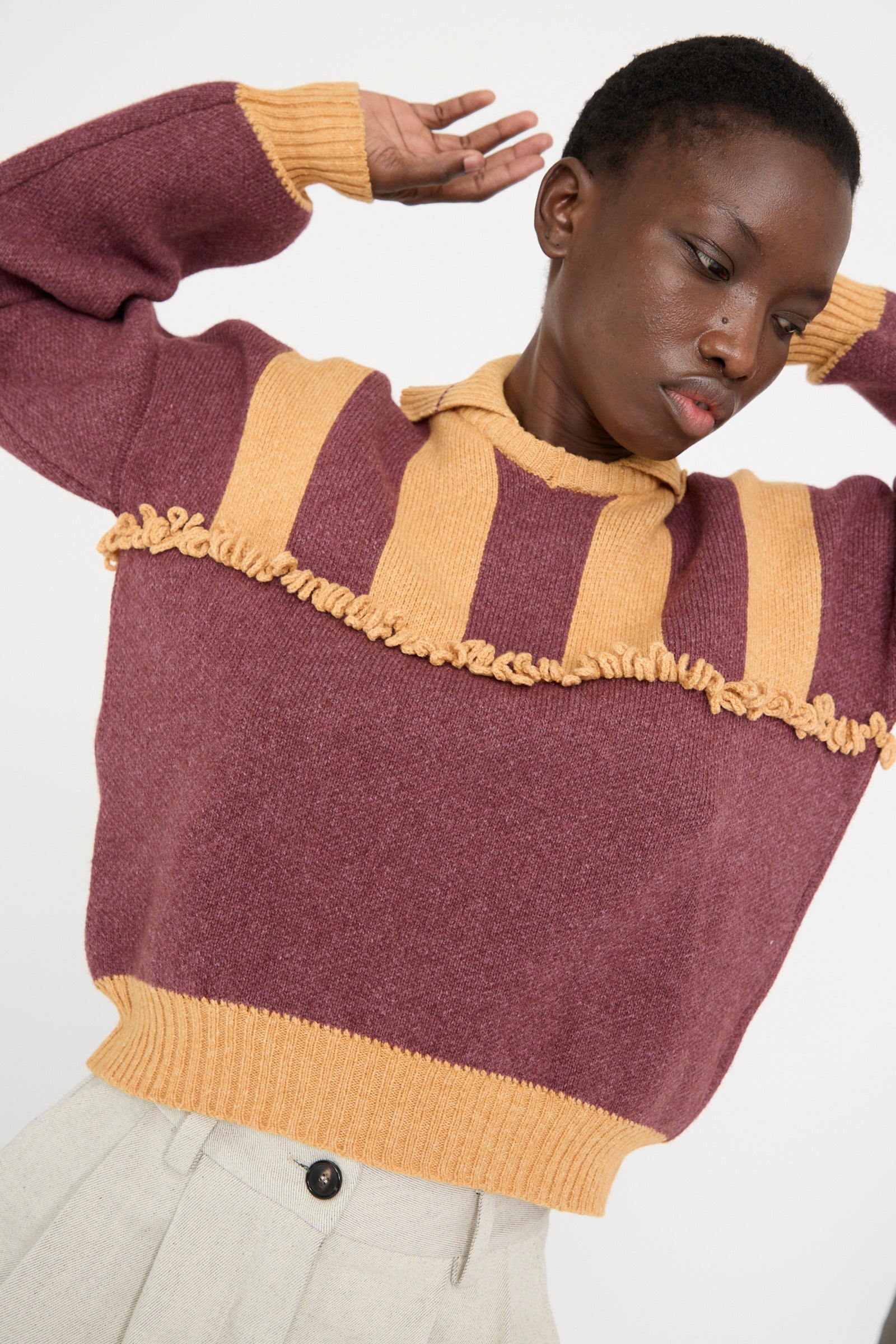 A model in a Cawley British Wool and Cotton Striped Amelia Pullover in Cassis and Aconite, featuring a striped yoke and ruffled design, exudes luxury with arms raised.