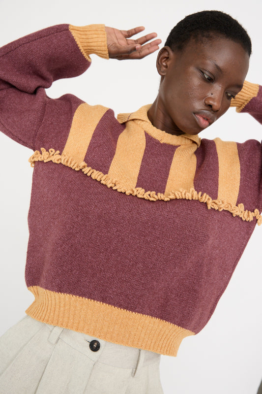 A model in a Cawley British Wool and Cotton Striped Amelia Pullover in Cassis and Aconite, featuring a striped yoke and ruffled design, with arms raised. 