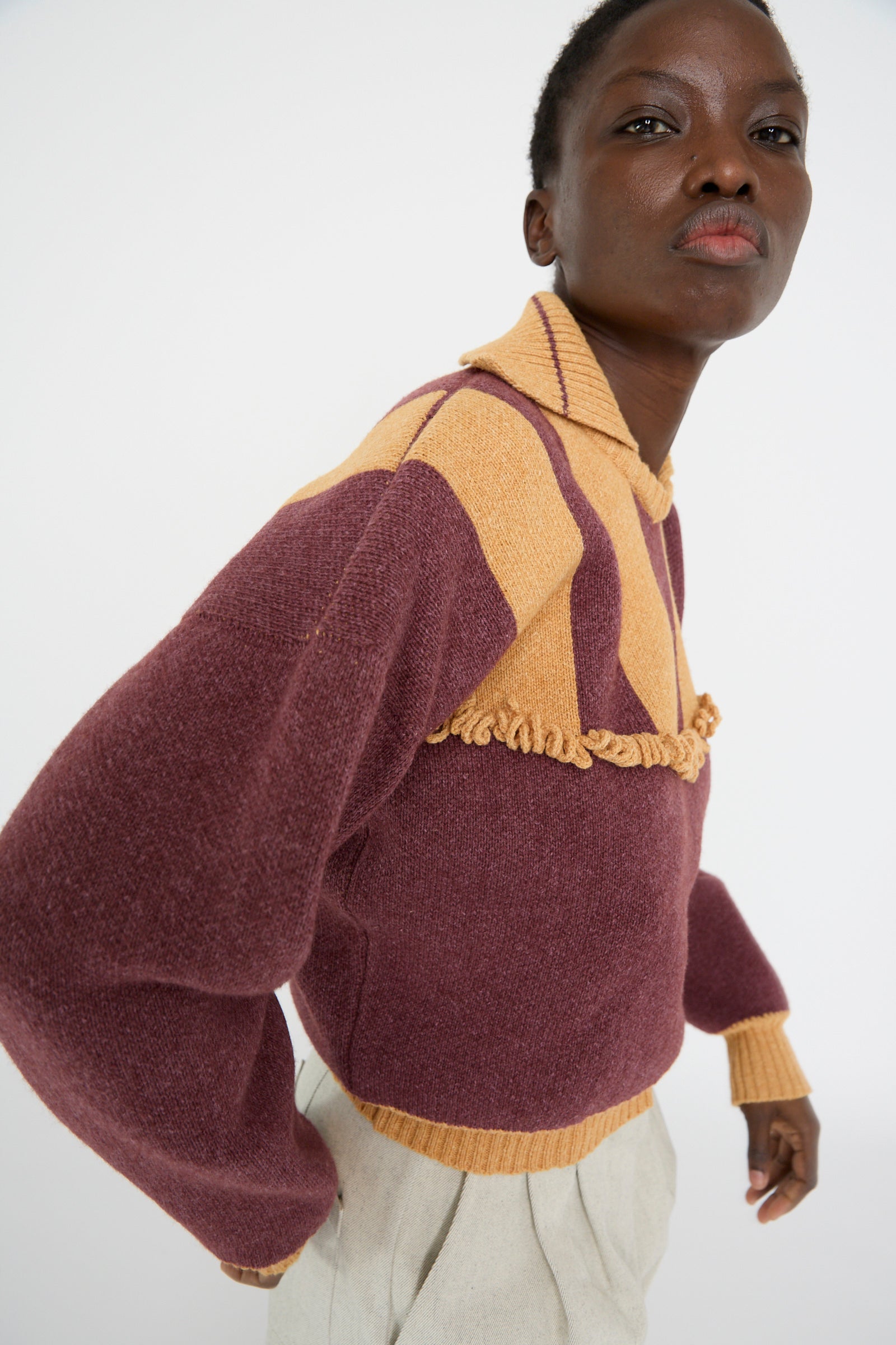 A person wearing the Cawley British Wool and Cotton Striped Amelia Pullover in Cassis and Aconite, paired with light-colored pants, stands against a white background.