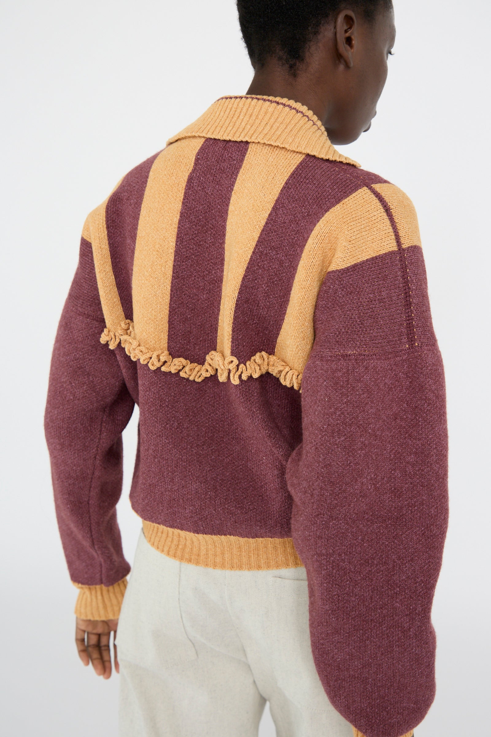 From behind, a person wears the Cawley British Wool and Cotton Striped Amelia Pullover in Cassis and Aconite. The sweater features multicolored knit, tan and purple stripes with ruffled details, paired elegantly with white pants for a cozy yet stylish look.