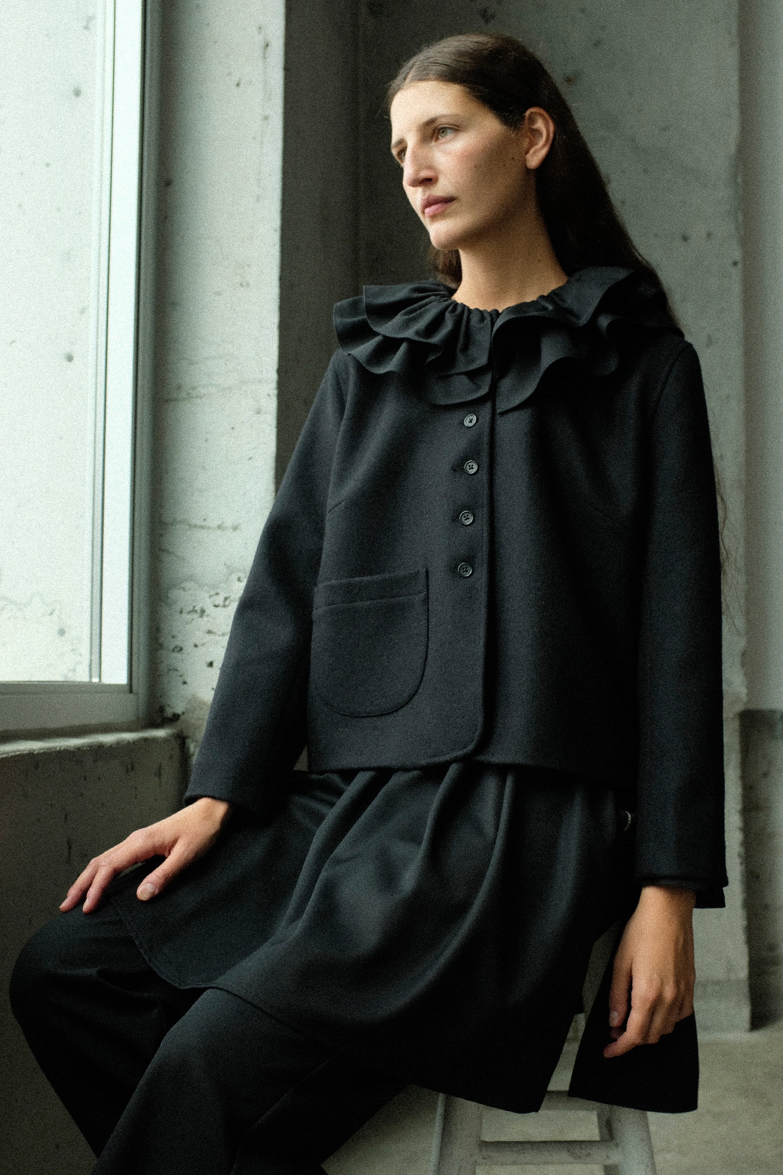 A woman seated indoors wearing Cawley's British Wool Rowan Jacket in Black, and British Lightweight Wool Layered Skirt Trouser in Black, posed against a concrete wall near a window.