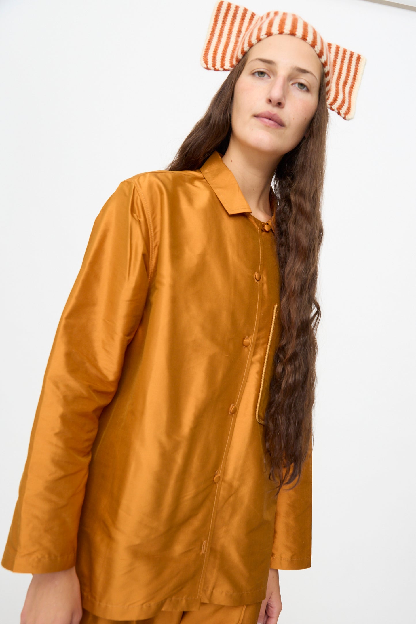 A person with long hair wearing a shiny burnt orange button-up top and matching pants, styled with the Cawley Brushed British Wool Stripe Bow Hat in Natural and Autumn, stands against a plain white background.