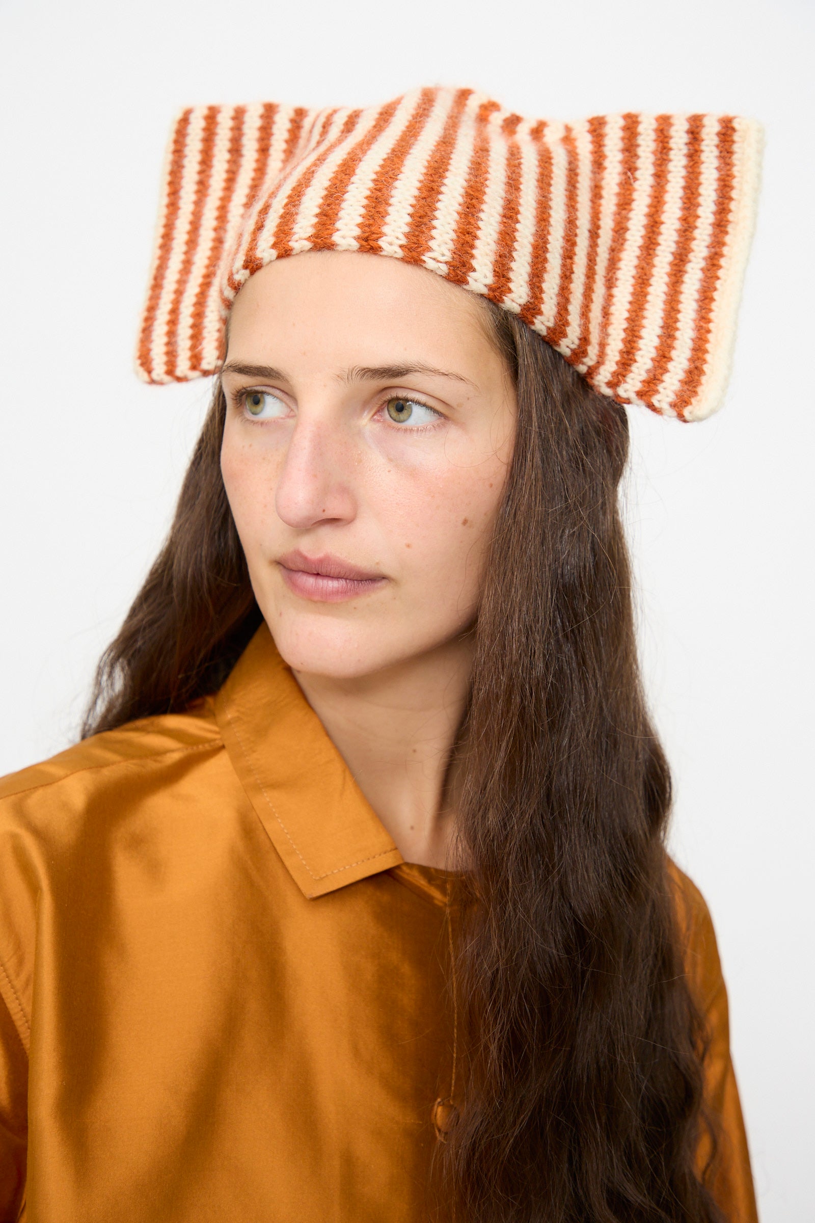 A person wears the Cawley Brushed British Wool Stripe Bow Hat in Natural and Autumn, featuring red-and-white stripes, paired with a rust-colored shirt and gazing slightly to the side against a plain background.