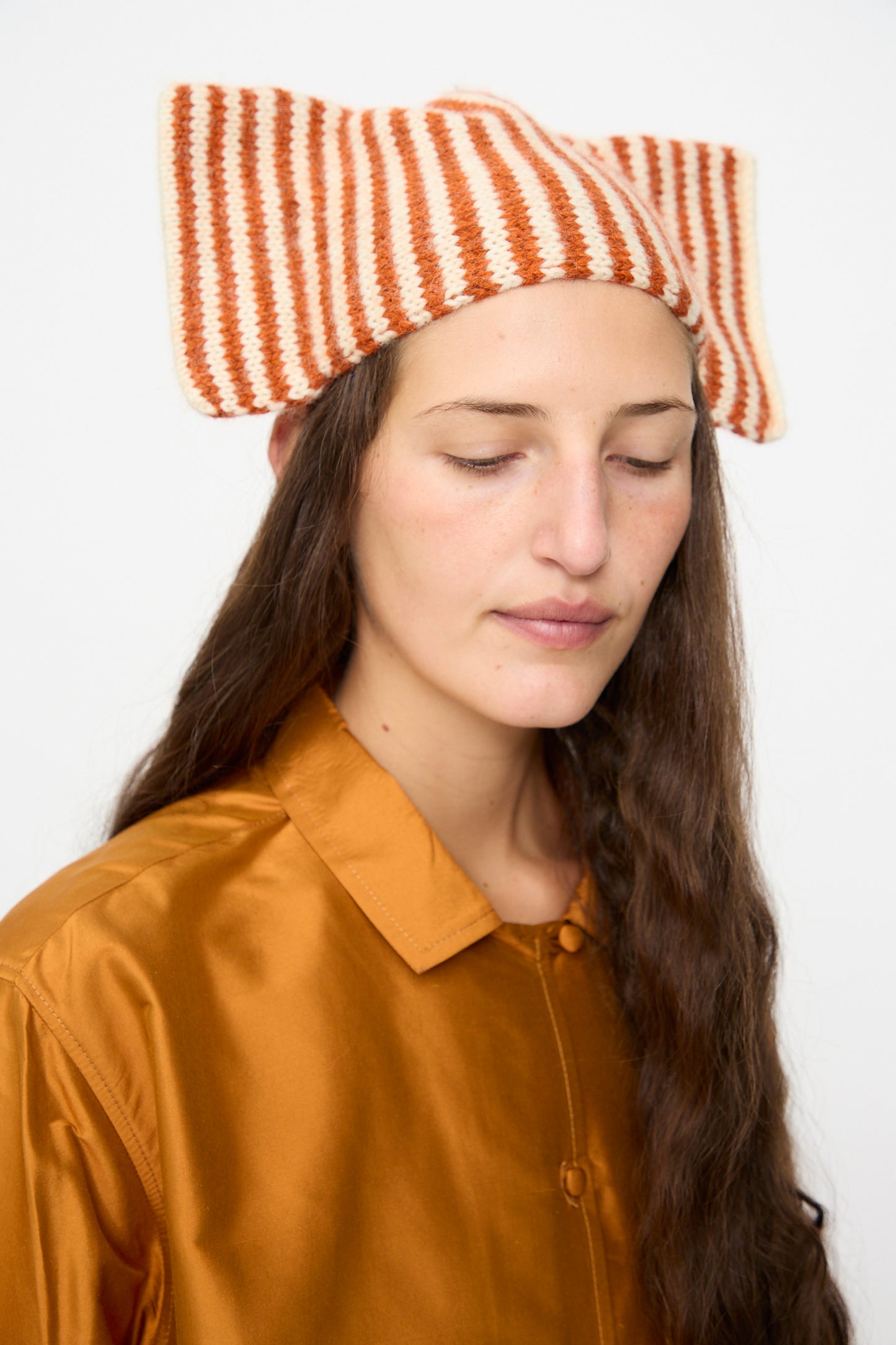 A person with long brown hair and closed eyes wears the Cawley Brushed British Wool Stripe Bow Hat in Natural and Autumn, paired with a shiny orange button-up shirt, against a plain white background.