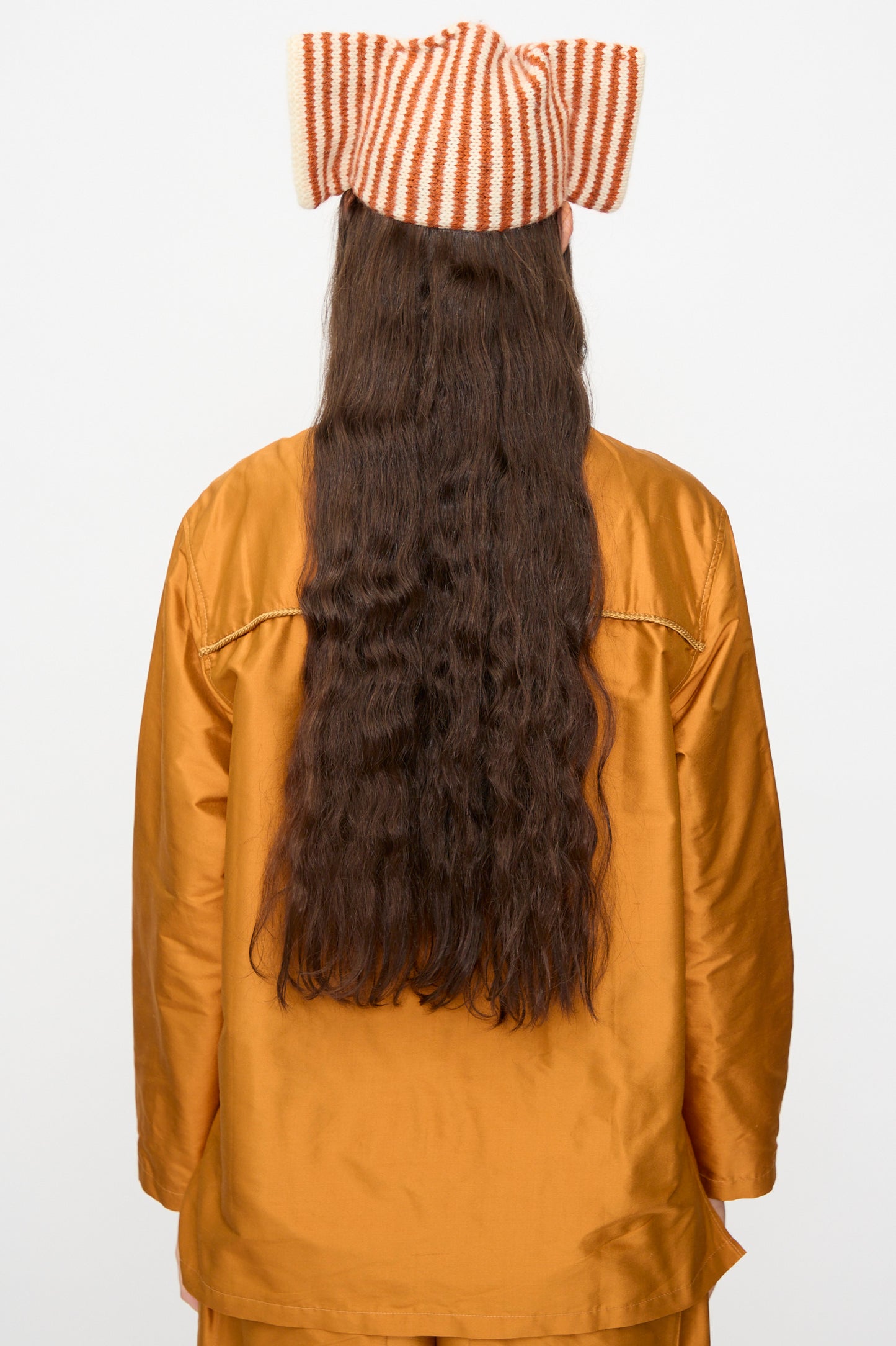 A person with long, wavy brown hair is seen from behind against a plain white background, wearing a gold-colored outfit and the Cawley Brushed British Wool Stripe Bow Hat in Natural and Autumn.