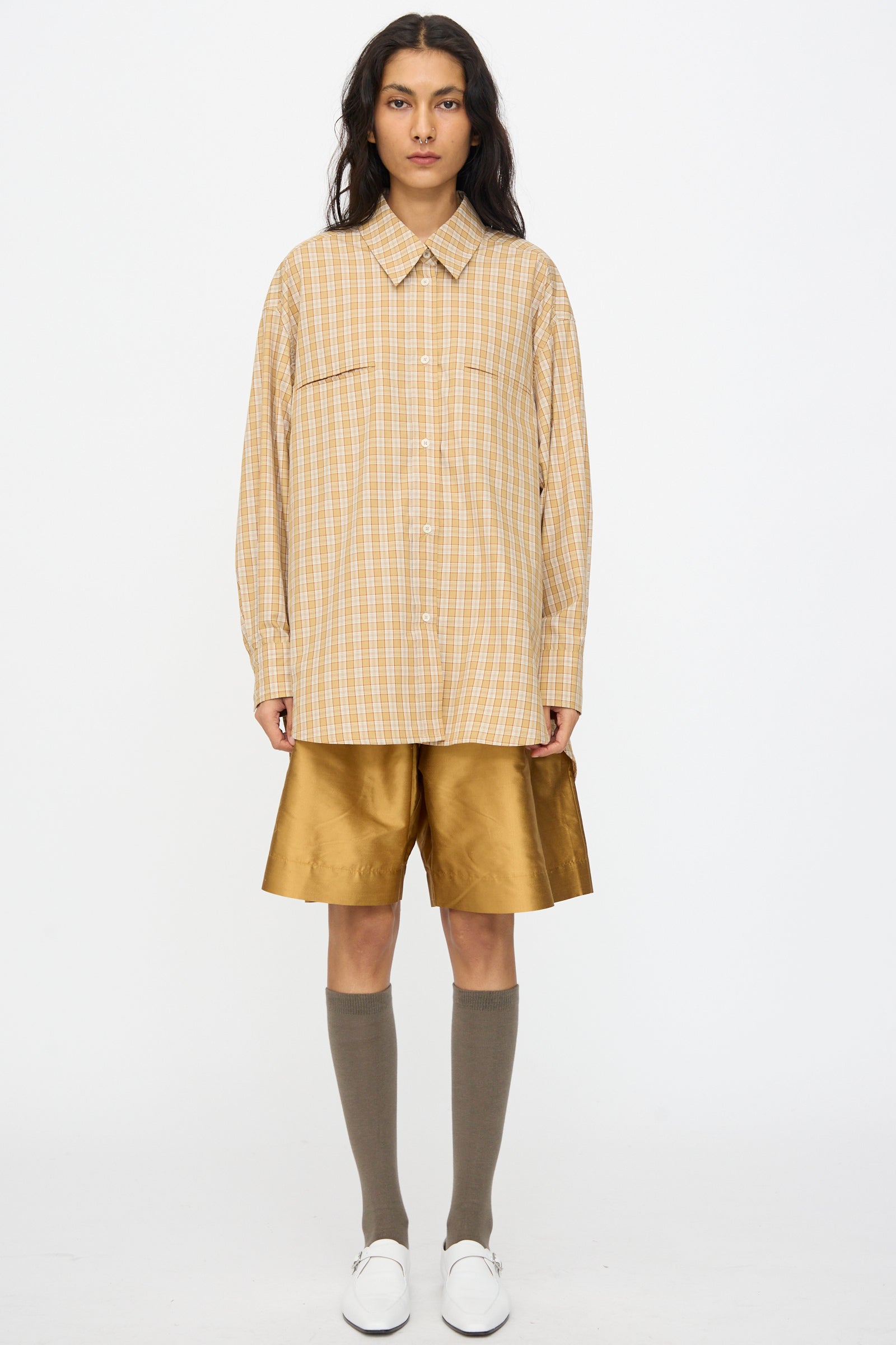 A person wears the Cawley Checked Cotton Phoebe Shirt in ochre, red, and purple check with gold knee-length shorts, gray knee-high socks, and white shoes, standing against a plain white background. The shirt is made in London.