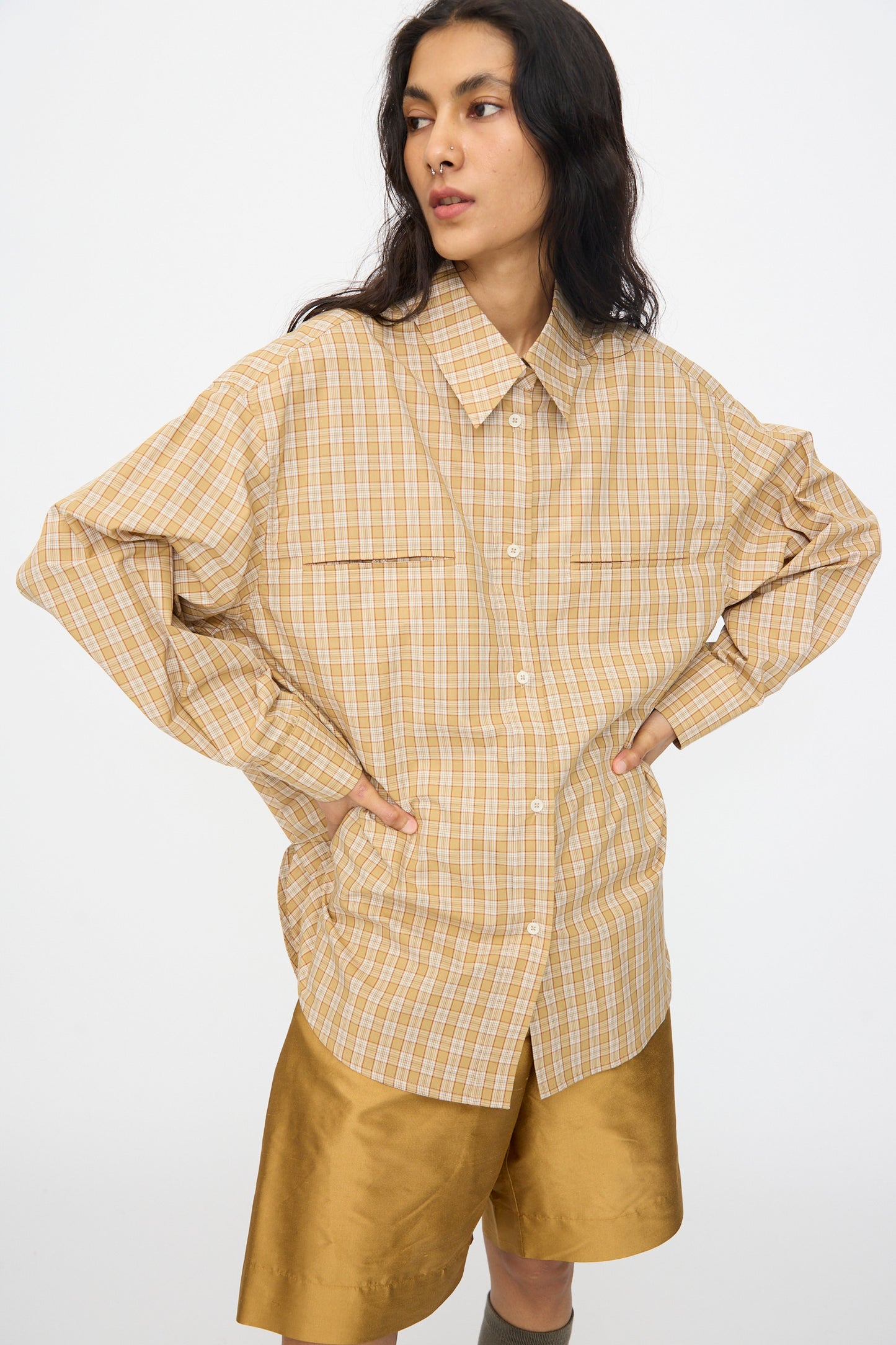 A person wears the Cawley Checked Cotton Phoebe Shirt in ochre, red, and purple check with gold satin shorts, made in London, standing with hands on hips against a plain white background.