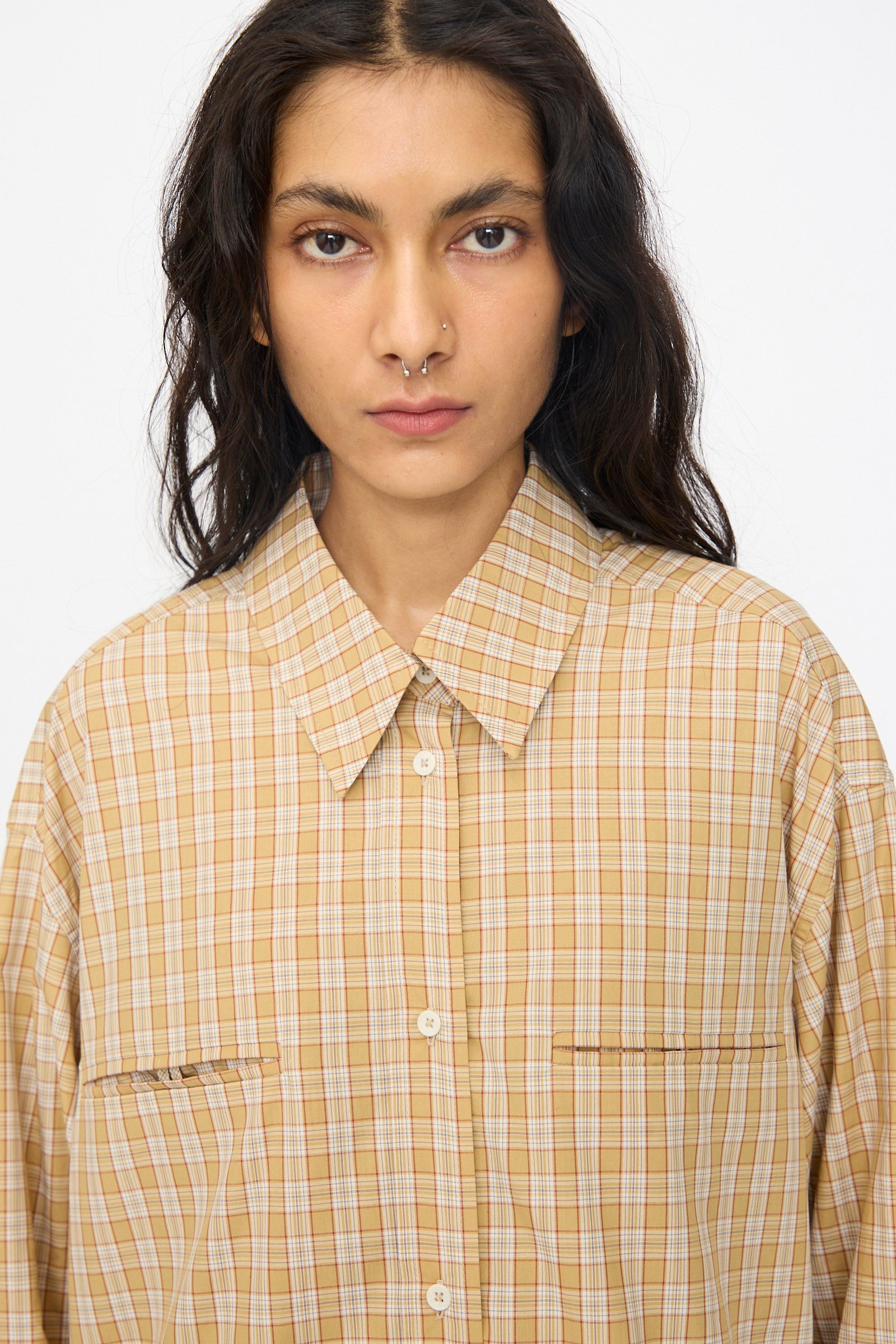 A person with long dark hair and a septum piercing wears the Cawley Checked Cotton Phoebe Shirt in ochre, red, and purple check, facing the camera against a plain white background.