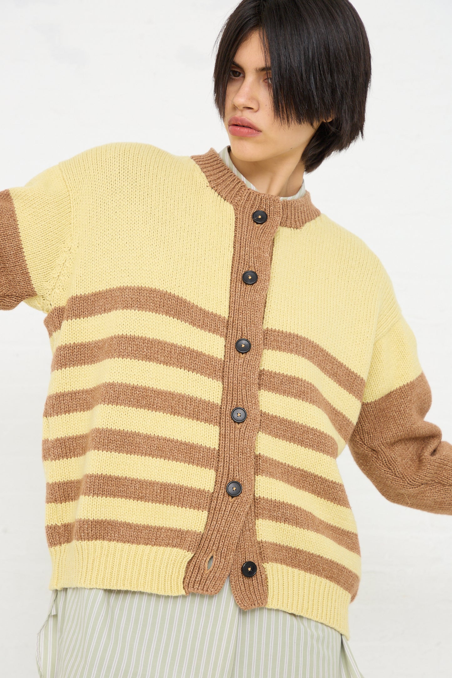 A person with a short dark hairstyle wearing a Cawley Cotton and Lambswool Textured Stripe Cardigan in Celery and Nutmeg over a light-colored vertical striped shirt, posing against a plain white background.