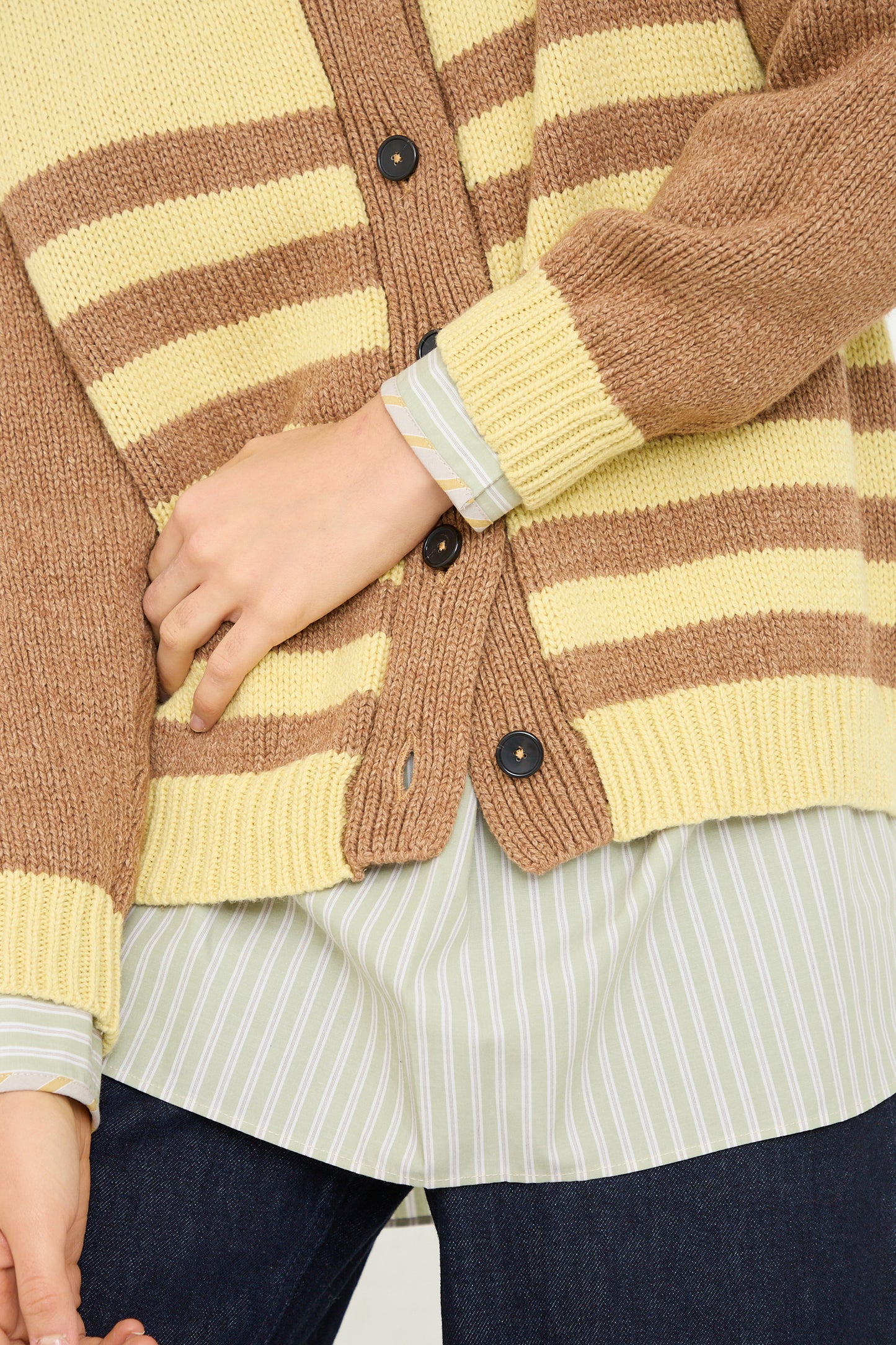 A close-up of a person wearing a Cawley Cotton and Lambswool Textured Stripe Cardigan in Celery and Nutmeg over a light blue and white striped Cotton shirt with their hand partially tucked into the front pocket of their outfit.