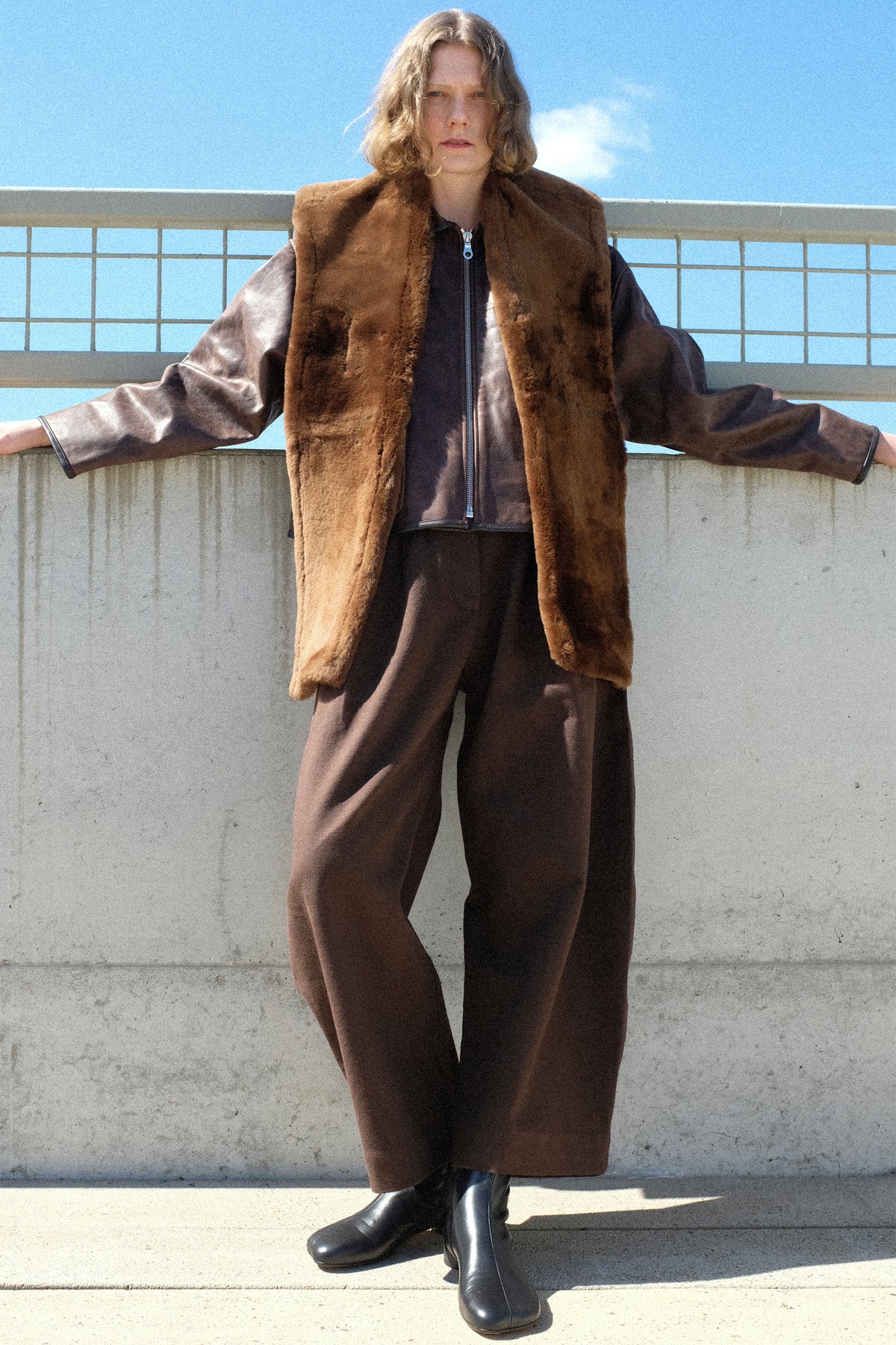 A woman standing outside against a concrete wall wearing Cawley's Leather Sheepskin Stole in Walnut, Waxed Leather Flying Jacket in Walnut, and the Japanese Gabardine Robyn Trouser in Espresso.