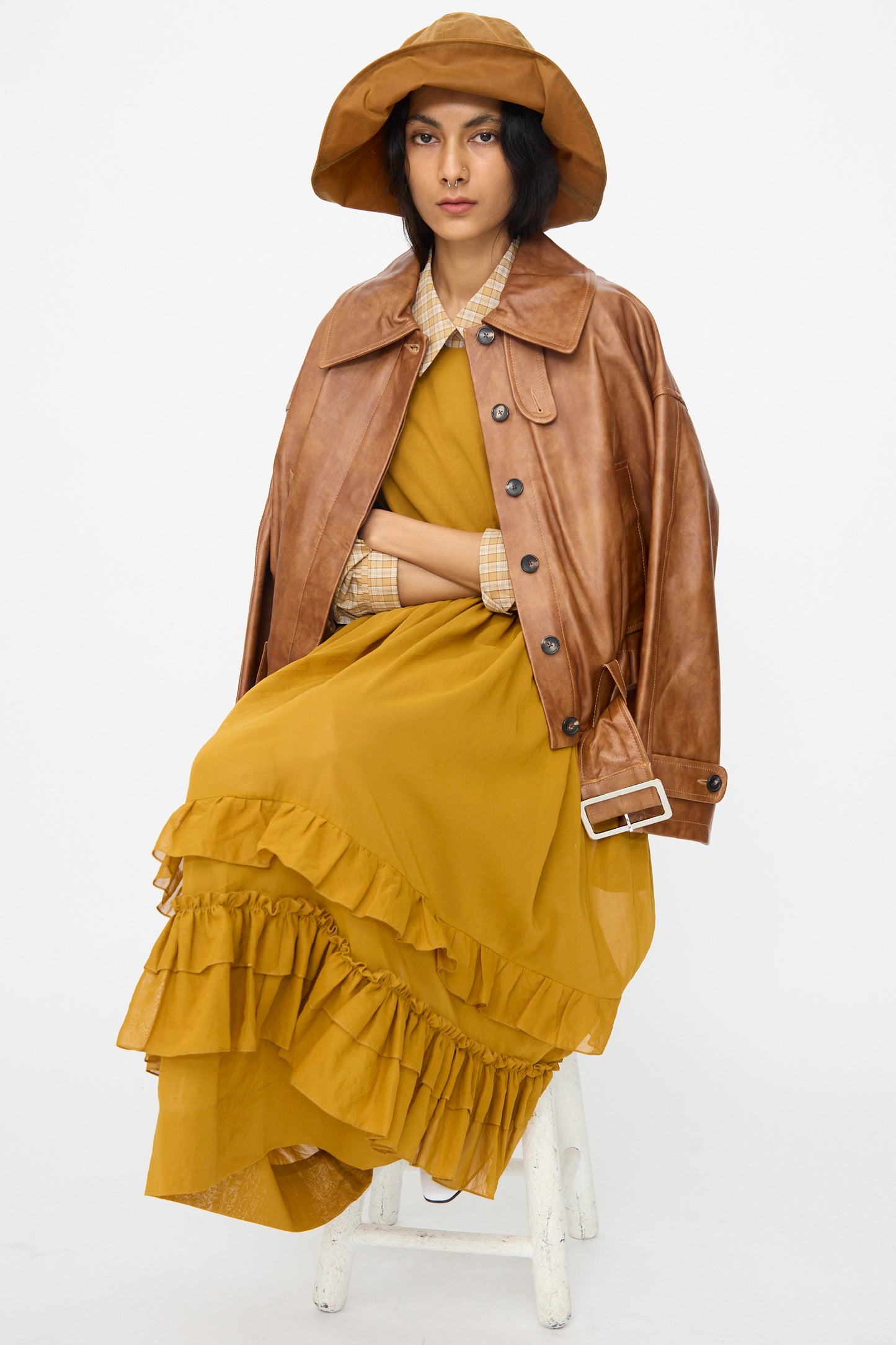 A person sits on a stool wearing the Cawley Holborn Leather Nara Jacket in Tan, a mustard yellow ruffled dress, plaid shirt, and brown wide-brimmed hat against a plain white background.
