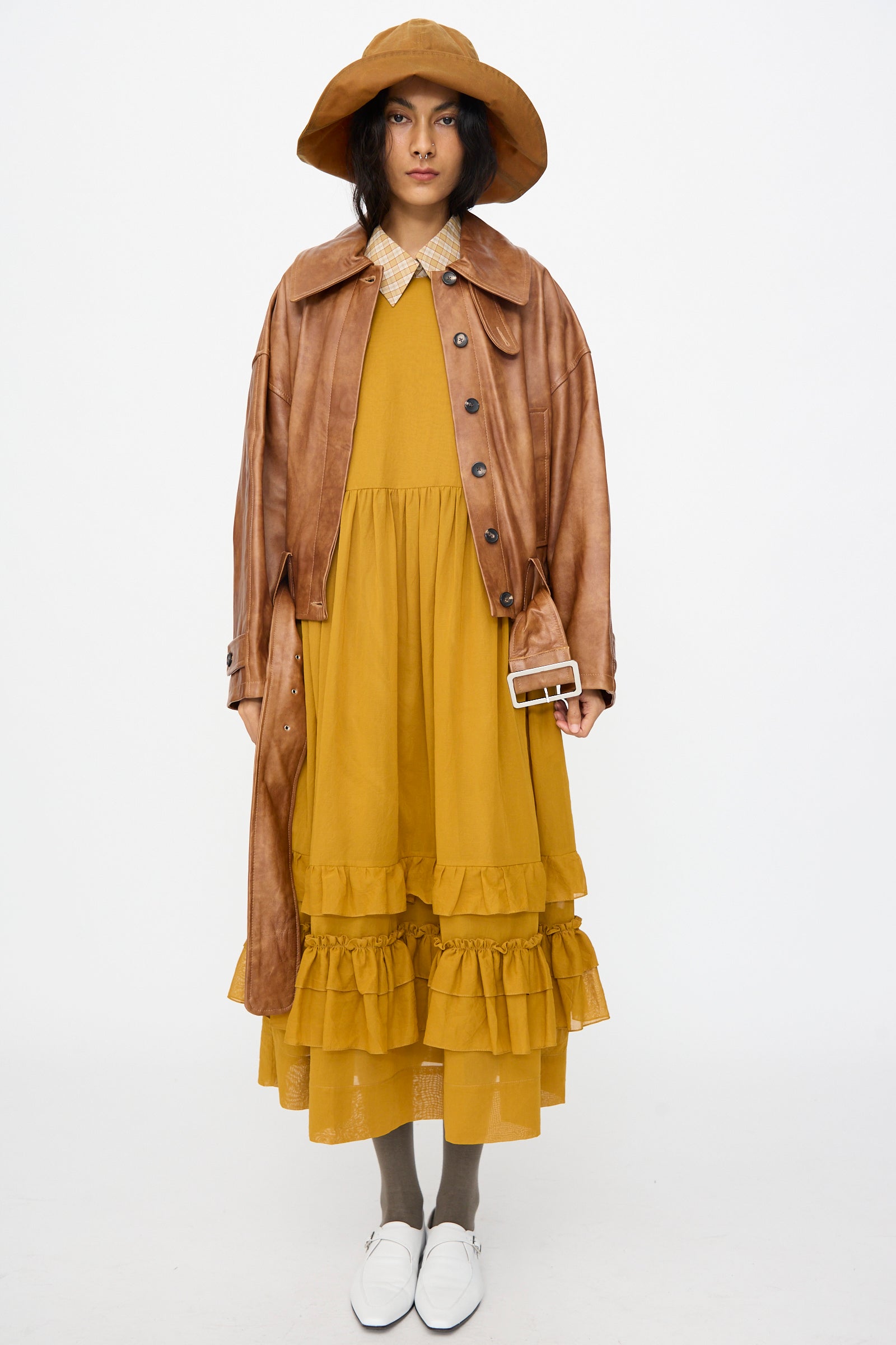 A person wearing a brown wide-brim hat, the Cawley Holborn Leather Nara Jacket in Tan, mustard yellow ruffled dress, gray socks, and white shoes stands against a plain white background.