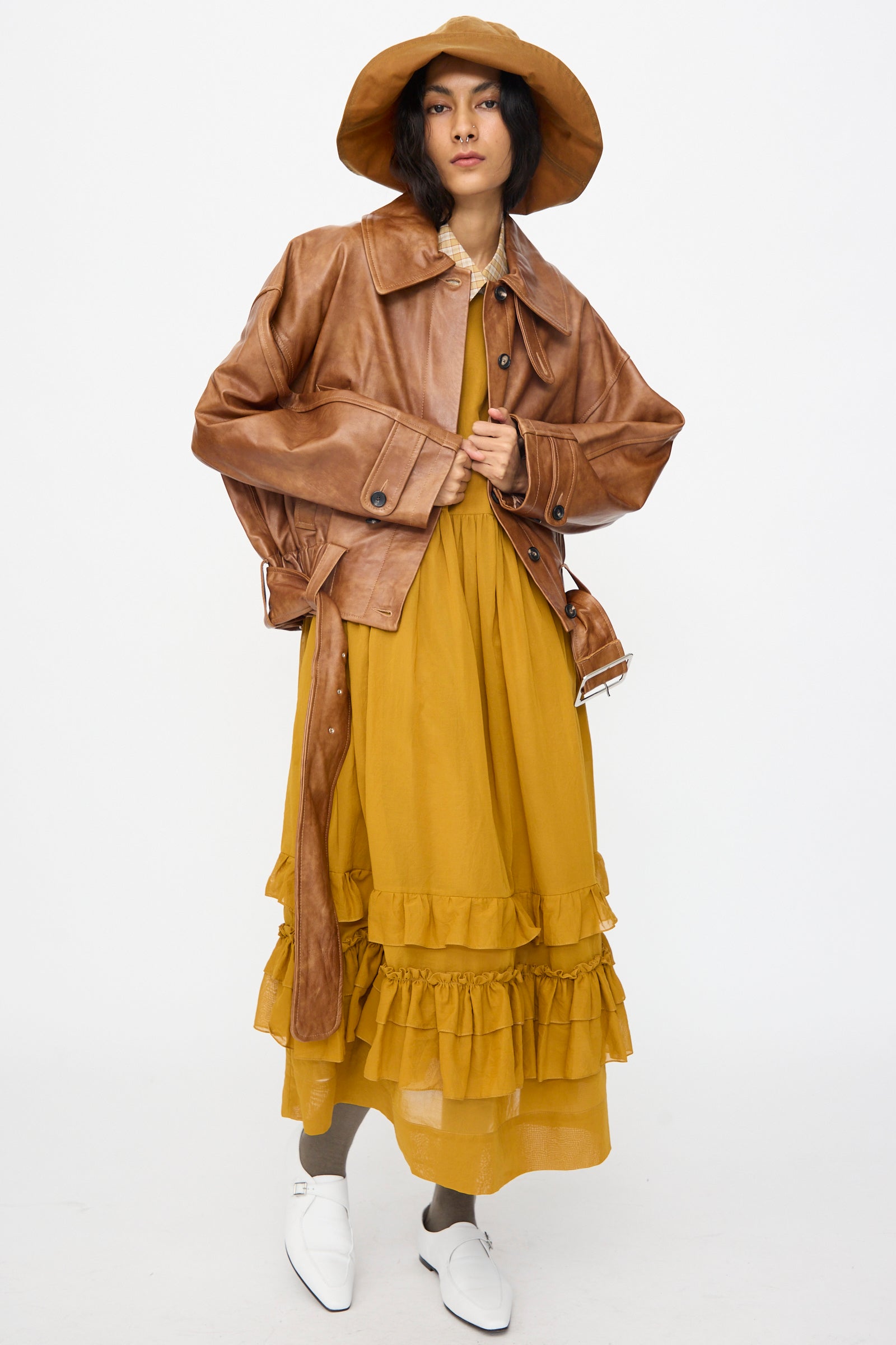 Individual clad in Cawley’s Holborn Leather Nara Jacket in Tan, a mustard yellow ruffled dress, gray socks, brown wide-brim hat, and white shoes stands against a plain white background.