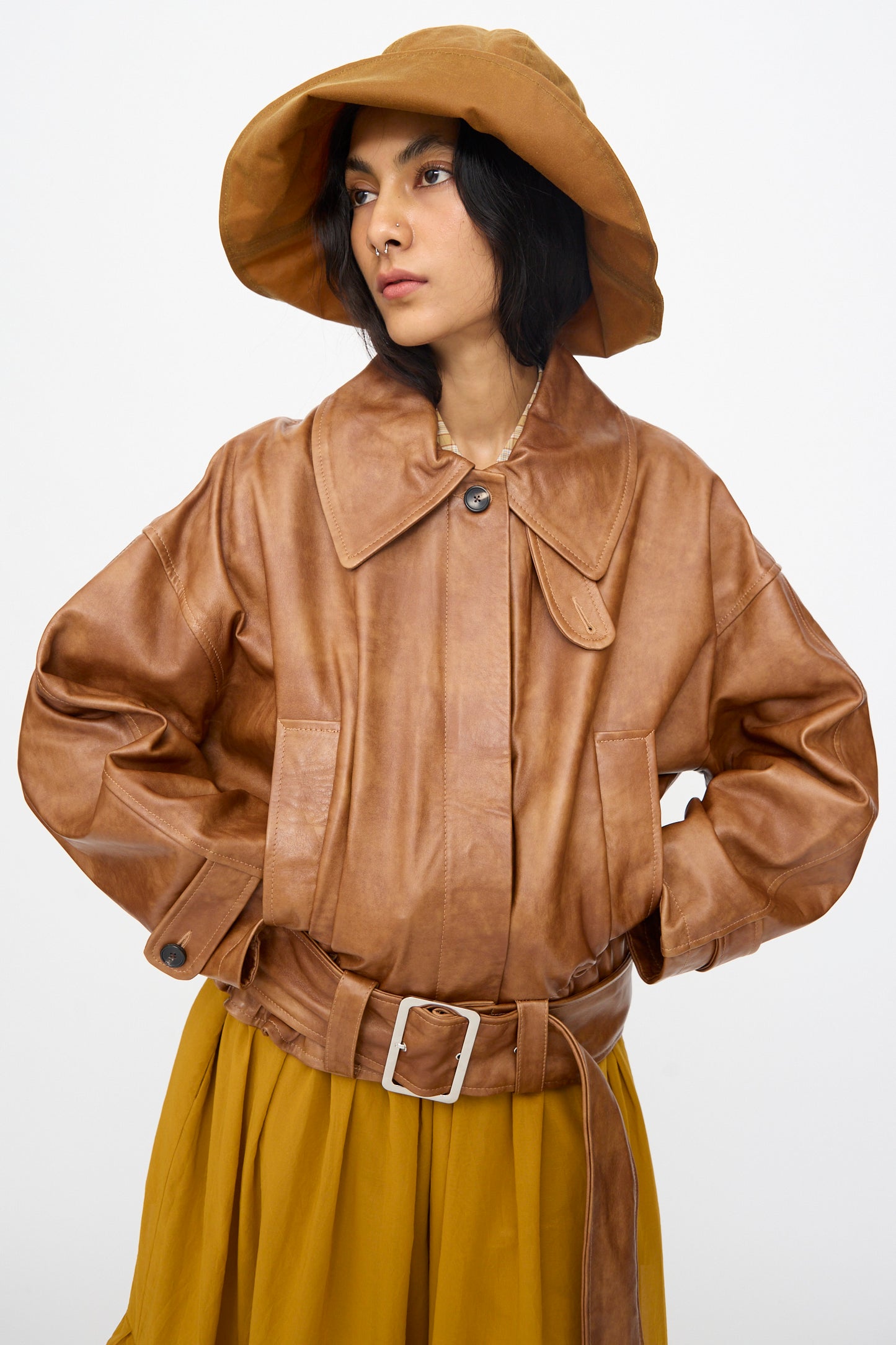 A person wears the Cawley Holborn Leather Nara Jacket in Tan with a wide collar and belt, paired with a brown wide-brim hat and mustard yellow skirt, standing against a plain white background.