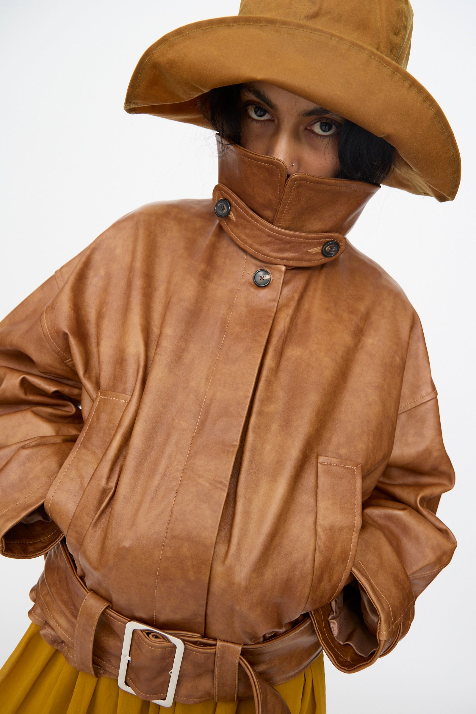 A woman in a wide-brimmed brown hat and Cawley's Holborn Leather Nara Jacket in Tan with a high collar looks directly at the camera against a plain white background.