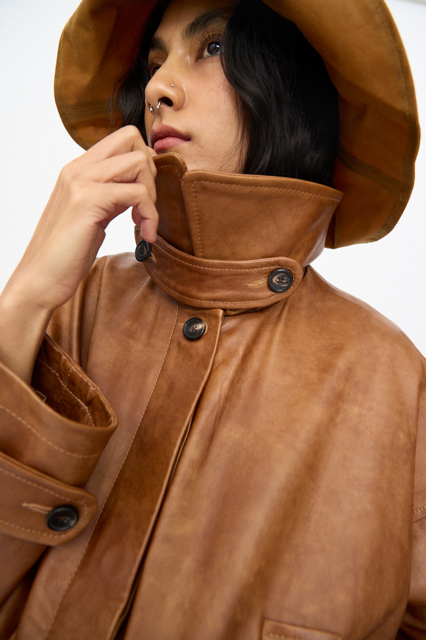 A person wears the Cawley Holborn Leather Nara Jacket in Tan with large buttons and a matching wide-brimmed hat, looking upward with a neutral expression. 