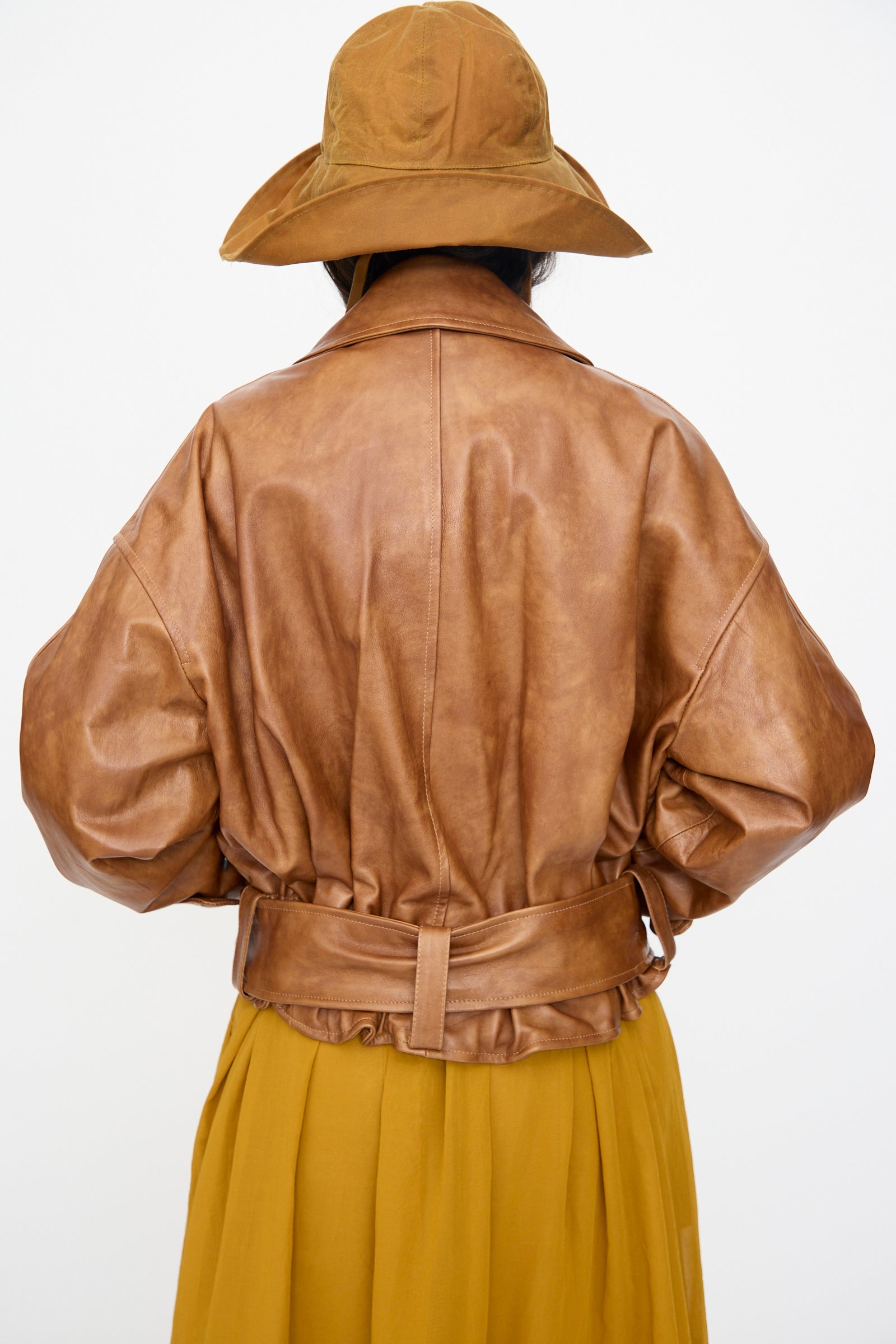 Person facing away, wearing Cawley’s Holborn Leather Nara Jacket in Tan with belt, yellow skirt, and wide-brimmed brown hat against a plain white background. Jacket is made in London.