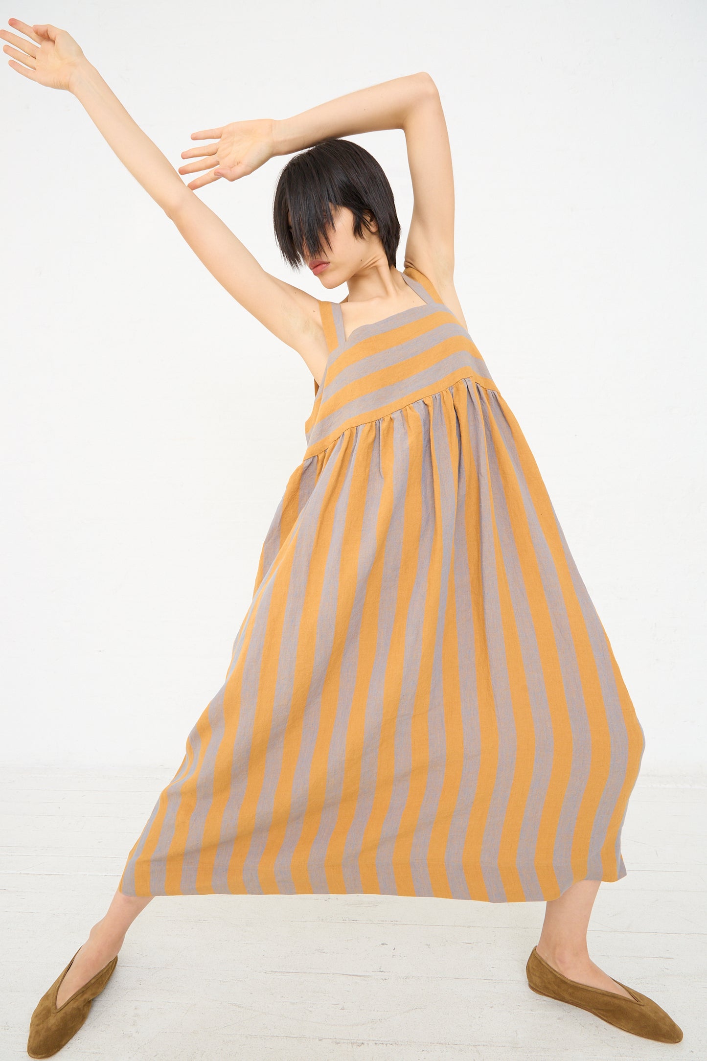 A woman in an oversized, striped Irish Linen Elba Dress in Bronze by Cawley is striking an expressive dance pose against a white background.