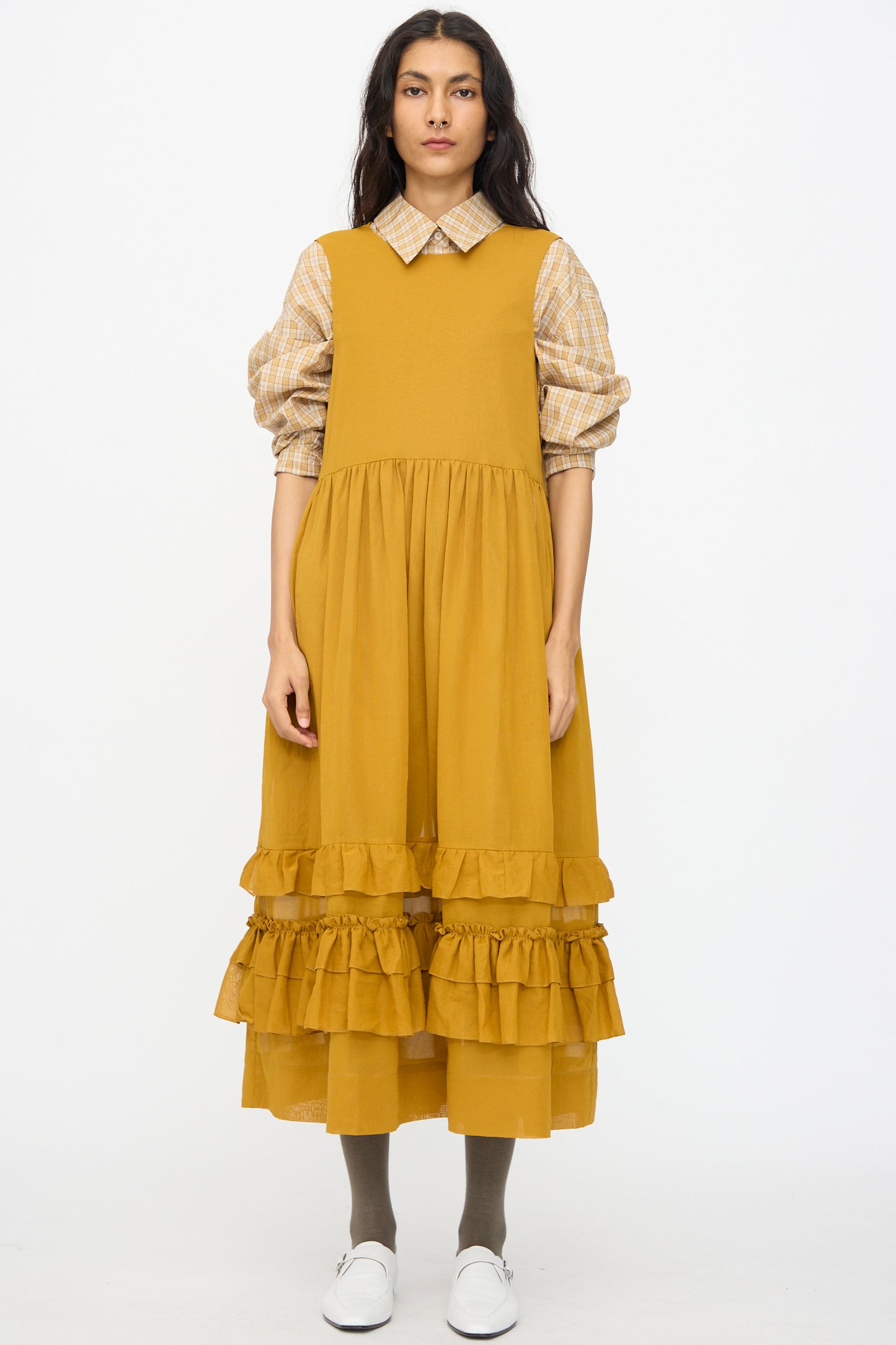 A woman stands facing forward in a Cawley Japanese Cotton Dyed Voile Genevieve Dress in Mustard, layered over a plaid shirt with gray tights and white shoes, set against a plain white background.