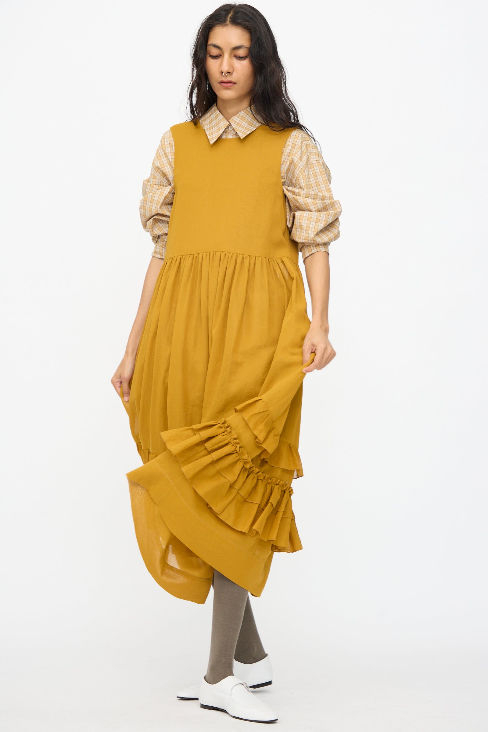 A woman wears the Cawley Japanese Cotton Dyed Voile Genevieve Dress in Mustard over a plaid shirt, with gray tights and white shoes, standing against a plain white background.