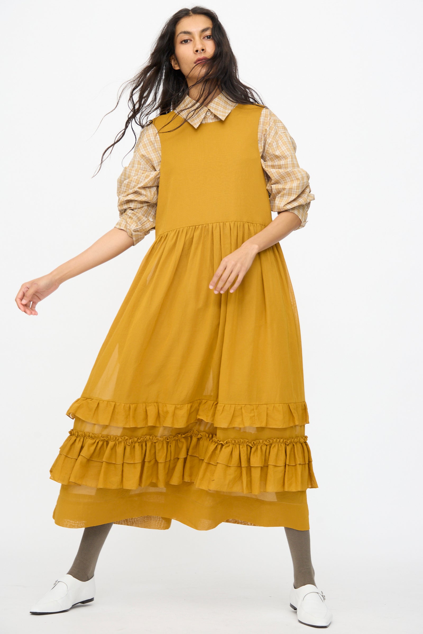 A person wears the Cawley Japanese Cotton Dyed Voile Genevieve Dress in Mustard over a plaid shirt, styled with gray tights and white shoes, standing against a plain white background.