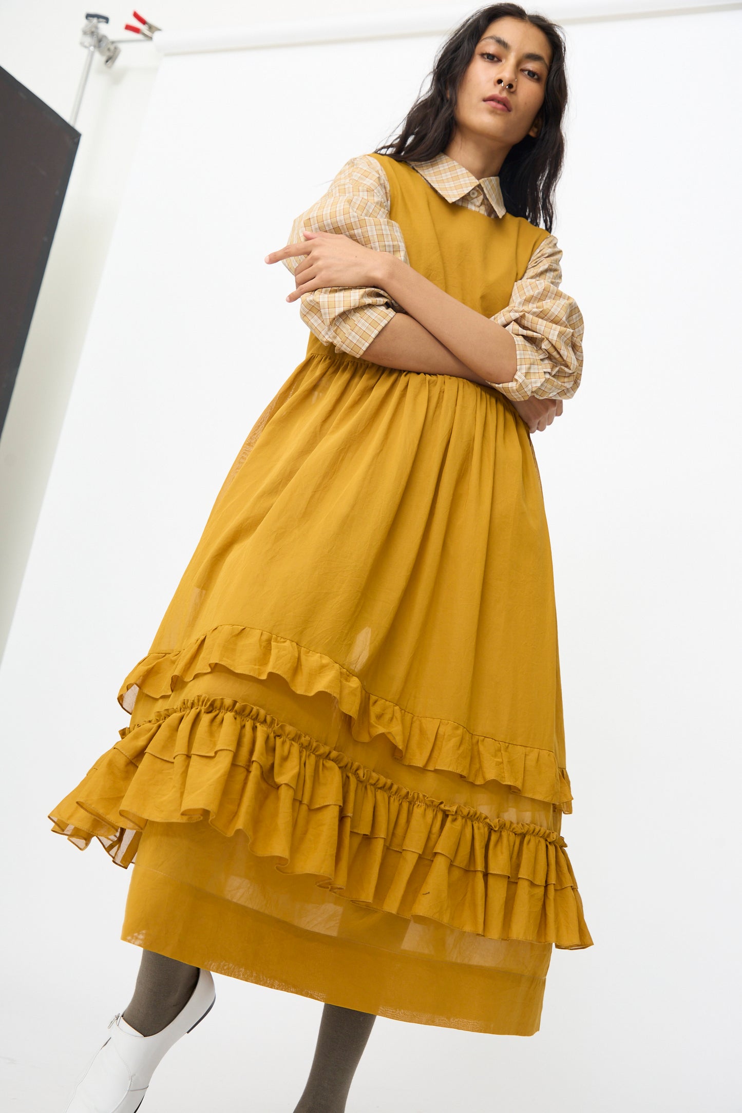 A woman wears the Cawley Japanese Cotton Dyed Voile Genevieve Dress in Mustard, styled over a plaid shirt, standing with arms crossed against a plain white background.