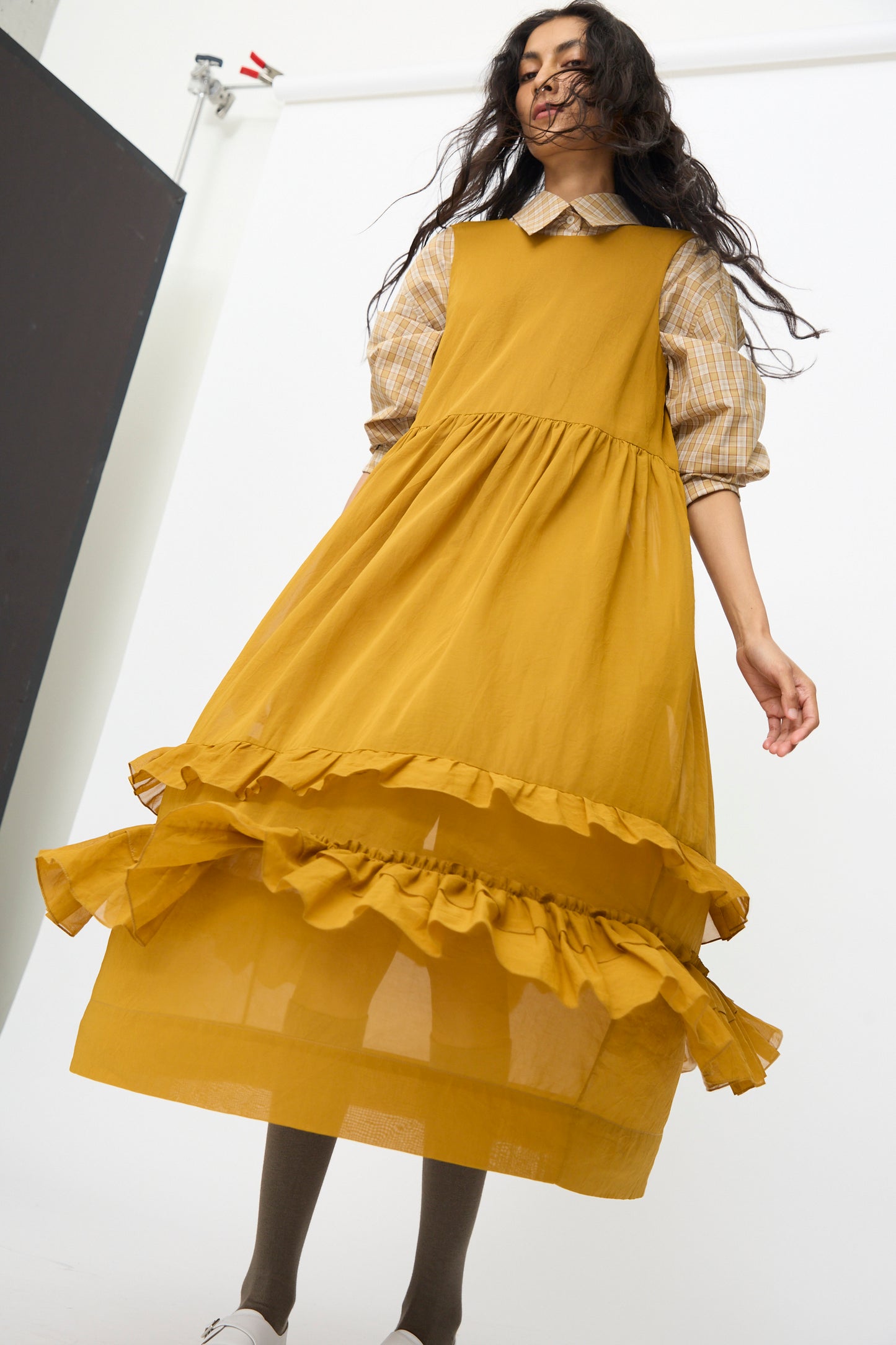 A woman in the Cawley Japanese Cotton Dyed Voile Genevieve Dress in Mustard, layered over a plaid shirt, poses before a white backdrop. The dress features a long, ruffled design and is captured from a low angle.
