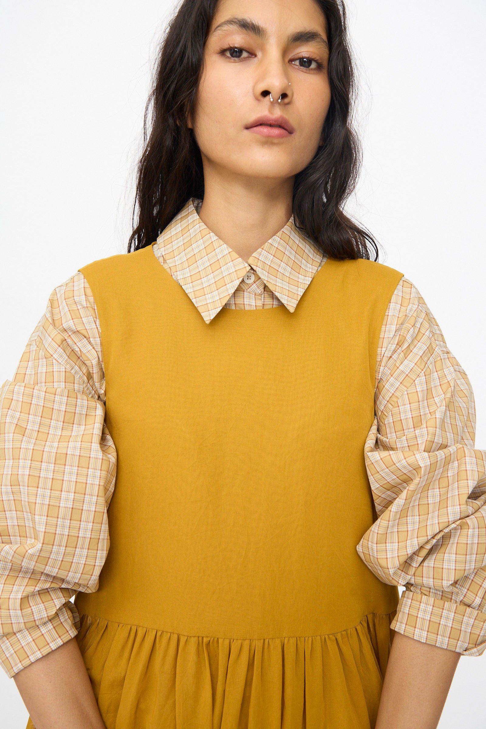 A woman wears the Cawley Japanese Cotton Dyed Voile Genevieve Dress in Mustard over a beige plaid puff-sleeve shirt, standing against a plain white background.
