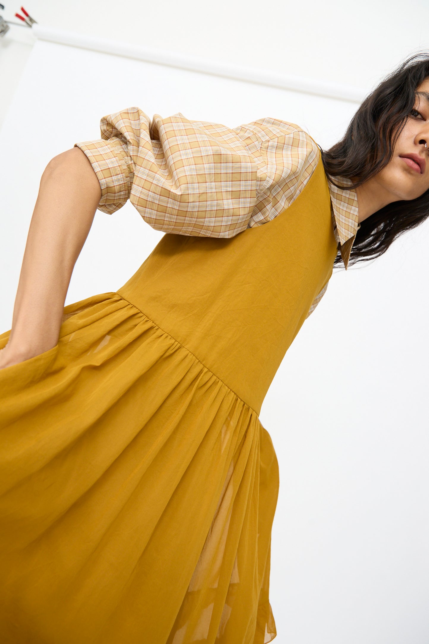 A woman wears the Cawley Japanese Cotton Dyed Voile Genevieve Dress in mustard, featuring plaid puff-sleeve detail, and poses against a plain white background.