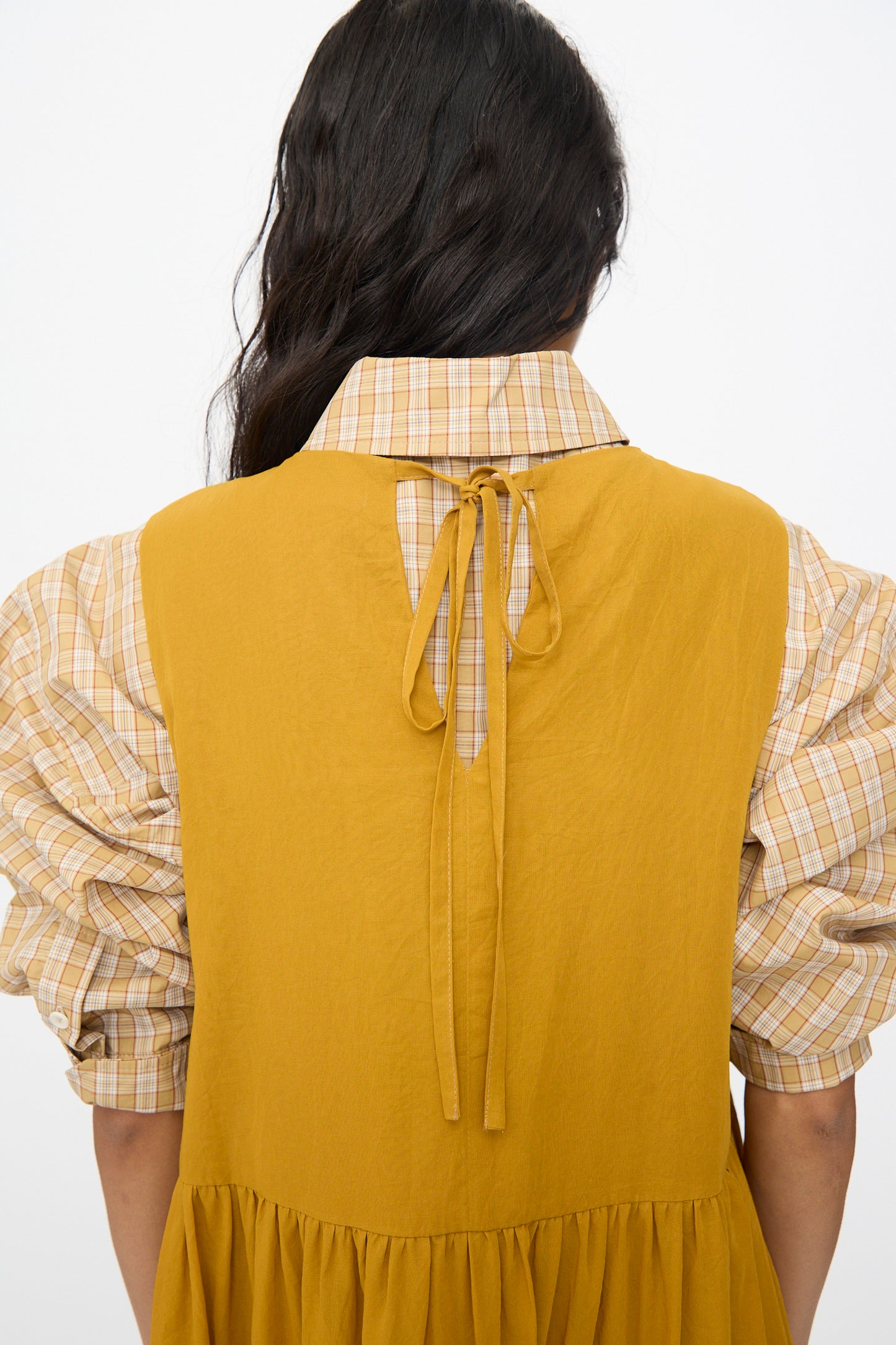 A woman with long, wavy dark hair is shown from the back wearing Cawley’s Japanese Cotton Dyed Voile Genevieve Dress in Mustard, featuring puffed plaid sleeves and a tie at the collar.
