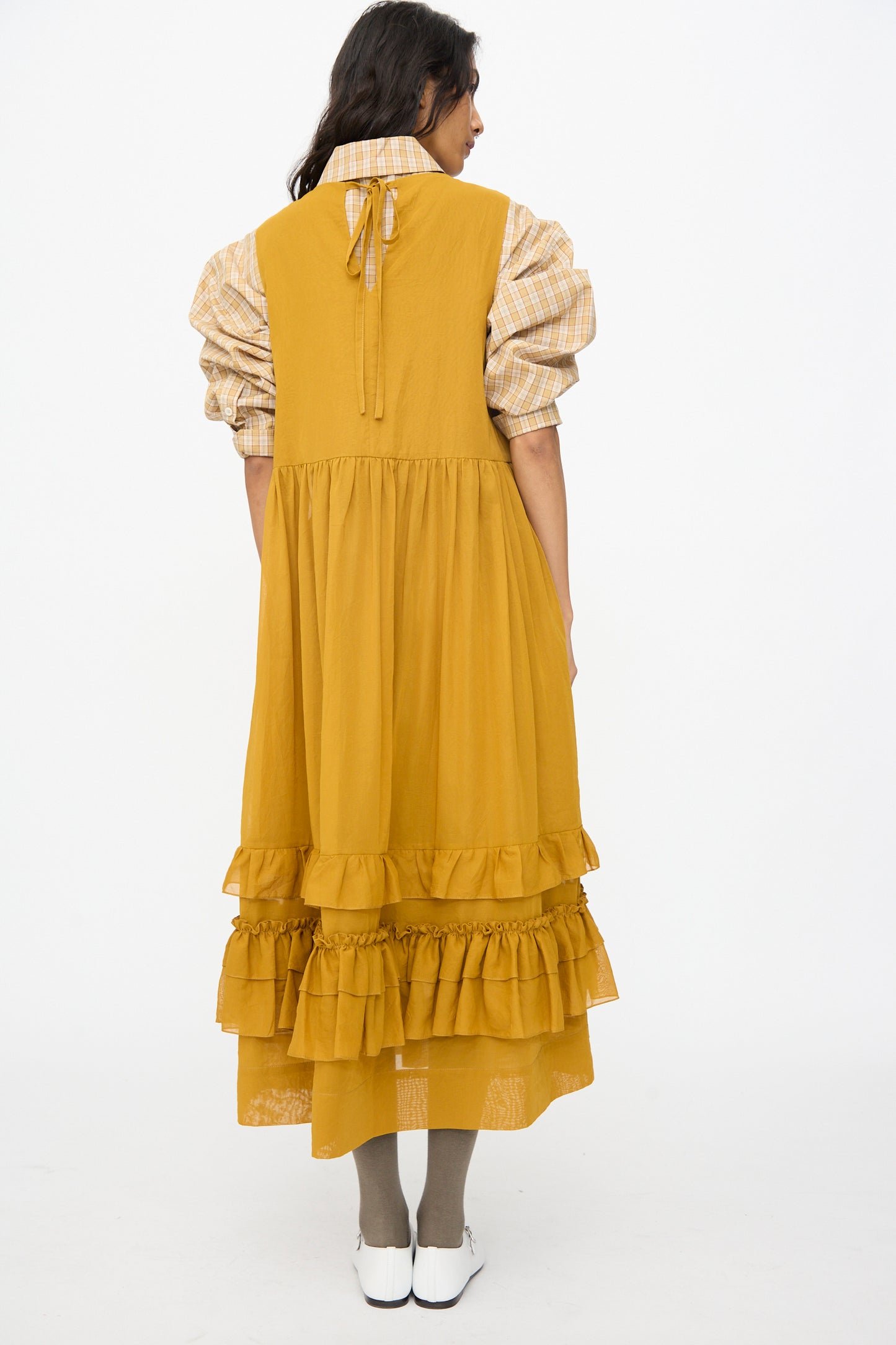 A woman stands against a white background, wearing the Cawley Japanese Cotton Dyed Voile Genevieve Dress in Mustard over a plaid puff-sleeve blouse, paired with beige tights and white shoes.