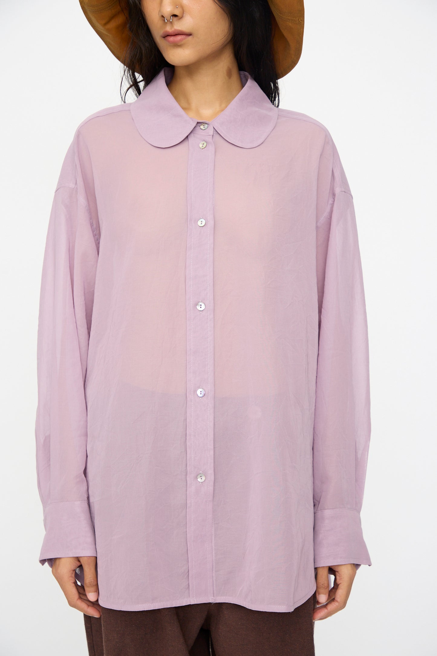 A person wears the Cawley Japanese Cotton Dyed Voile Phoebe Shirt in Purple, featuring a sheer finish and large rounded collar, paired with brown pants, set against a plain background.