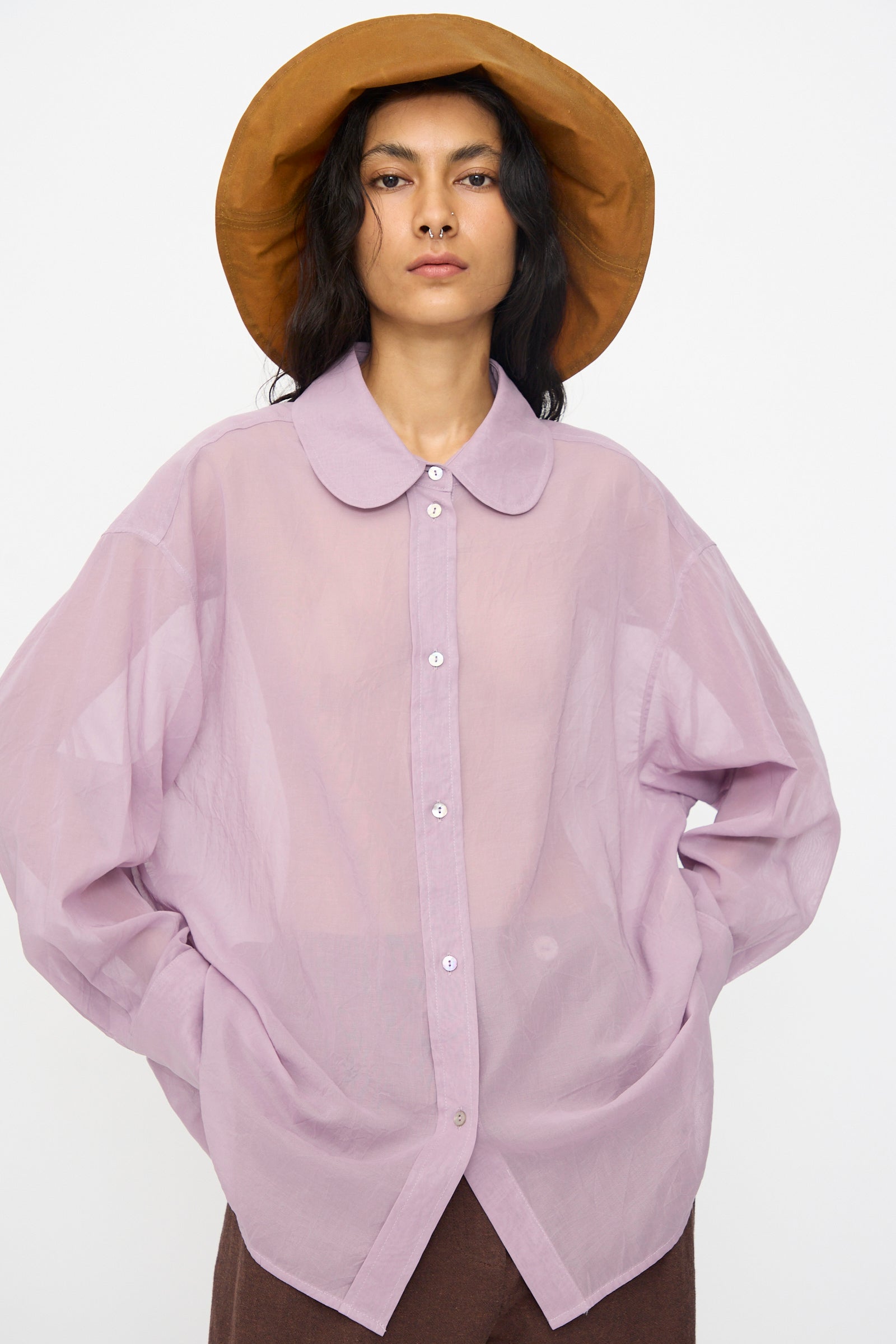 Person wearing the Cawley Japanese Cotton Dyed Voile Phoebe Shirt in Purple—an oversized fit, sheer button-up with a rounded collar—styled with a brown wide-brim hat and brown pants, standing against a plain white background.