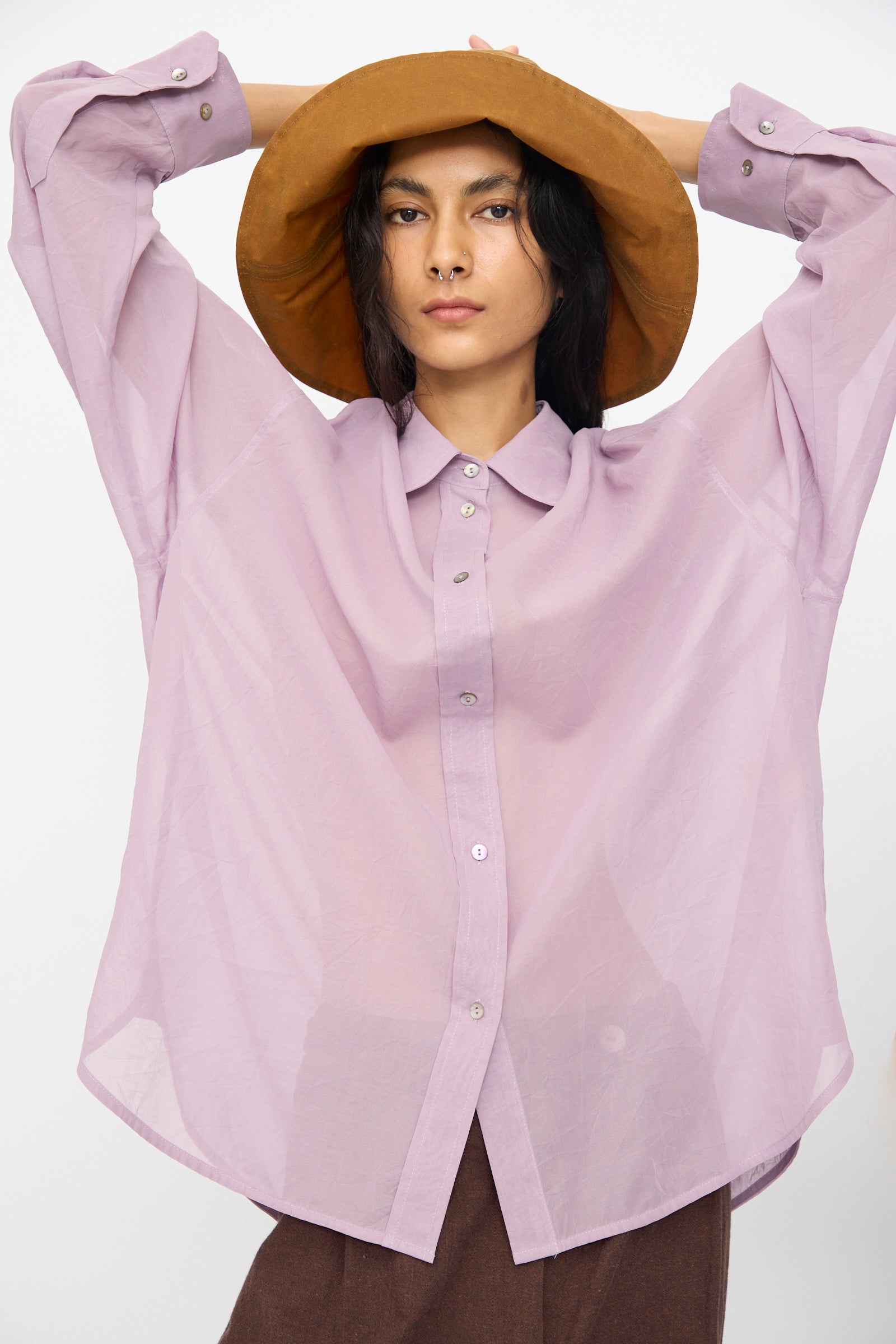 Person wearing the Cawley Japanese Cotton Dyed Voile Phoebe Shirt in Purple and a wide-brimmed brown hat, posing with arms raised behind their head against a plain background.
