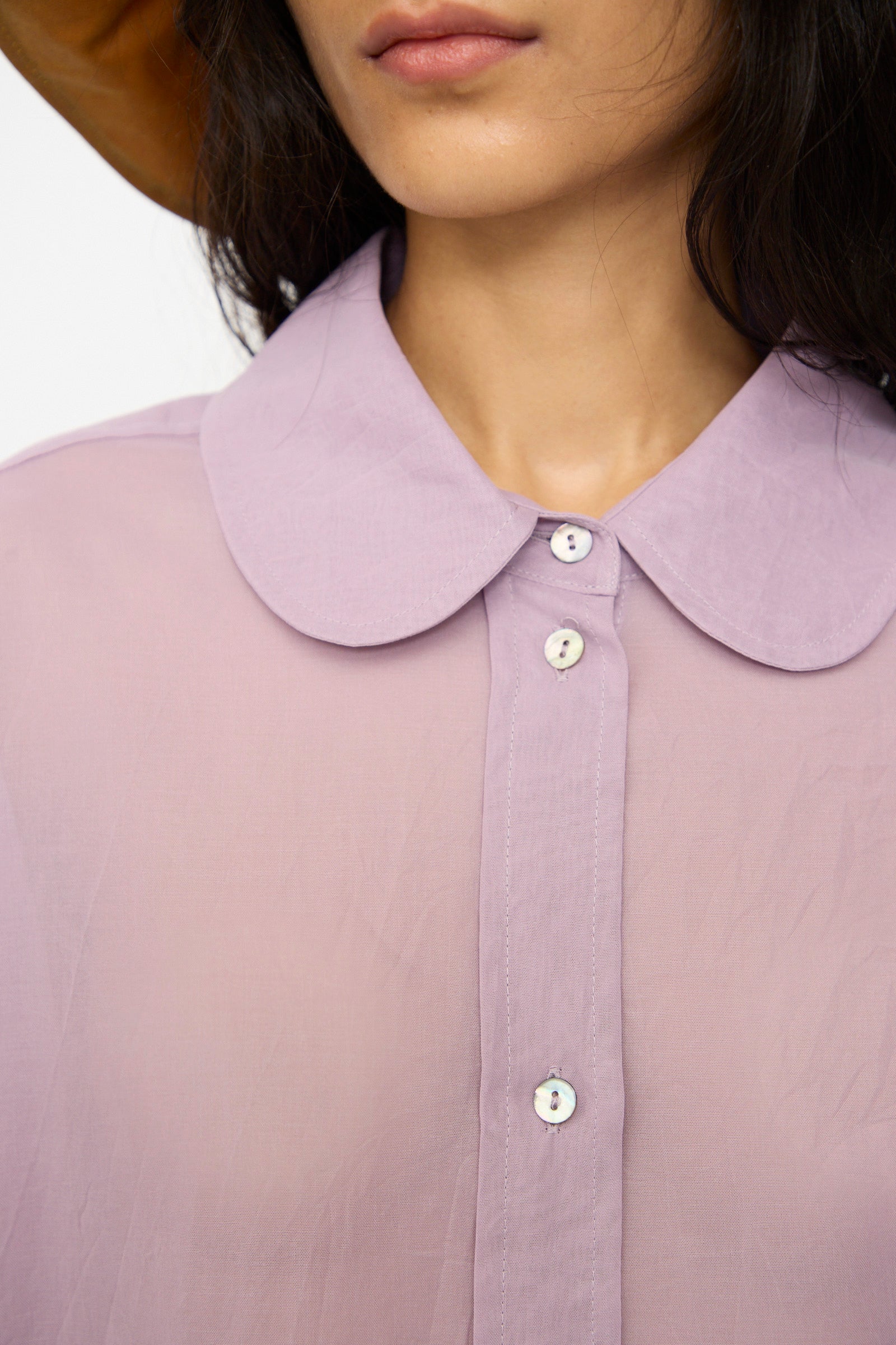 A woman wears the Cawley Japanese Cotton Dyed Voile Phoebe Shirt in Purple, an oversized sheer button-up with a rounded collar, paired with a light brown hat—shown from the shoulders up.