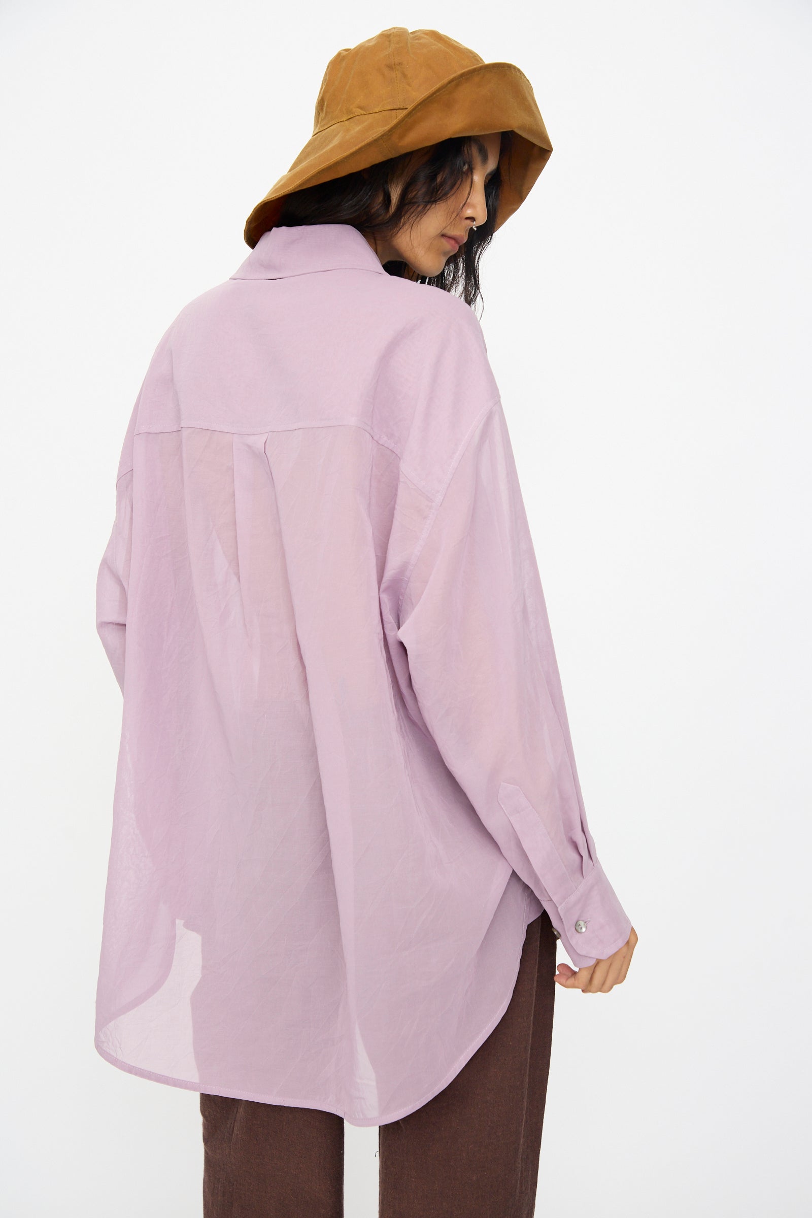 A person wears the Cawley Japanese Cotton Dyed Voile Phoebe Shirt in Purple, paired with brown pants and a brown bucket hat, standing with their back turned and looking over their shoulder against a white background.