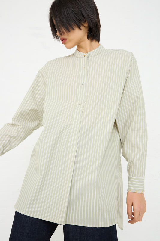 A woman in a vertically striped, light green and white Japanese Cotton Ines Shirt in Mint and Ecru by Cawley, with a high collar, partially unbuttoned, paired with dark blue jeans.